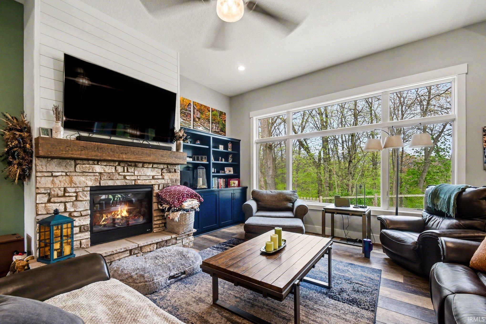 Living area featuring an expansive picture window, stone fireplace with a wood-beam mantel, built-in shelving, wood-finish flooring, and a ceiling fan