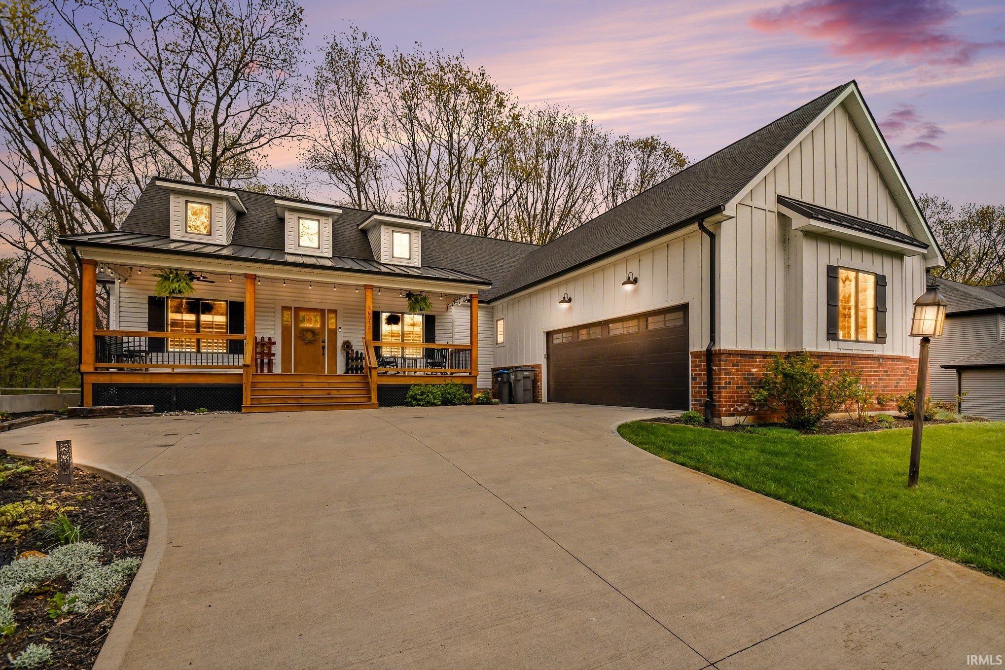 Contemporary farmhouse residence featuring a wide concrete driveway, an expansive covered front porch with dark railings and natural wood columns, and multiple dormer windows