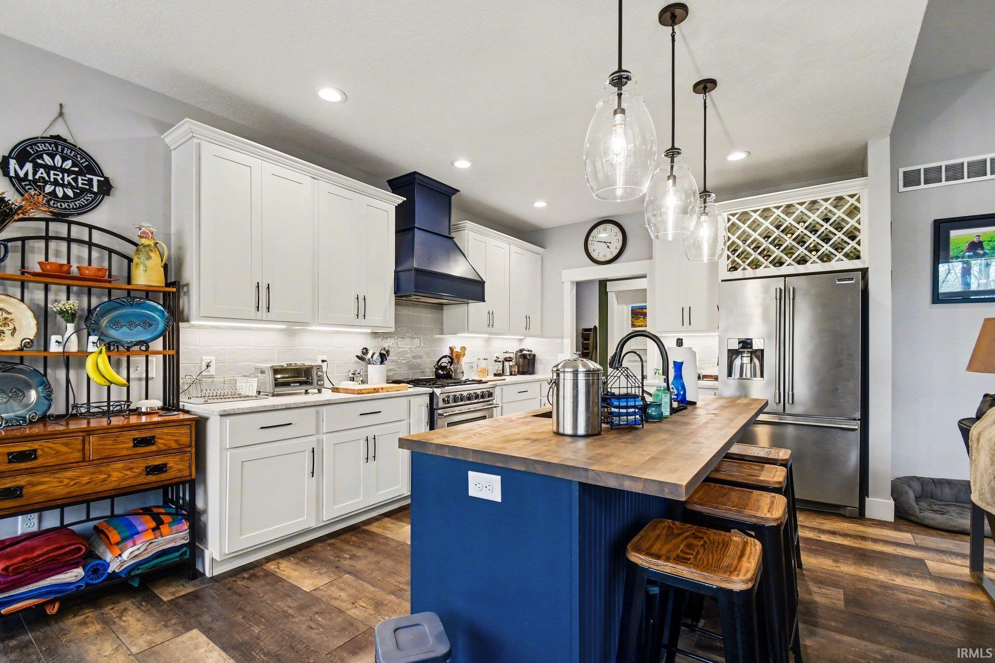 Open-concept kitchen featuring a central island with a wood-finish countertop, pendant lighting, shaker-style cabinetry, a professional-grade range, and built-in wine storage