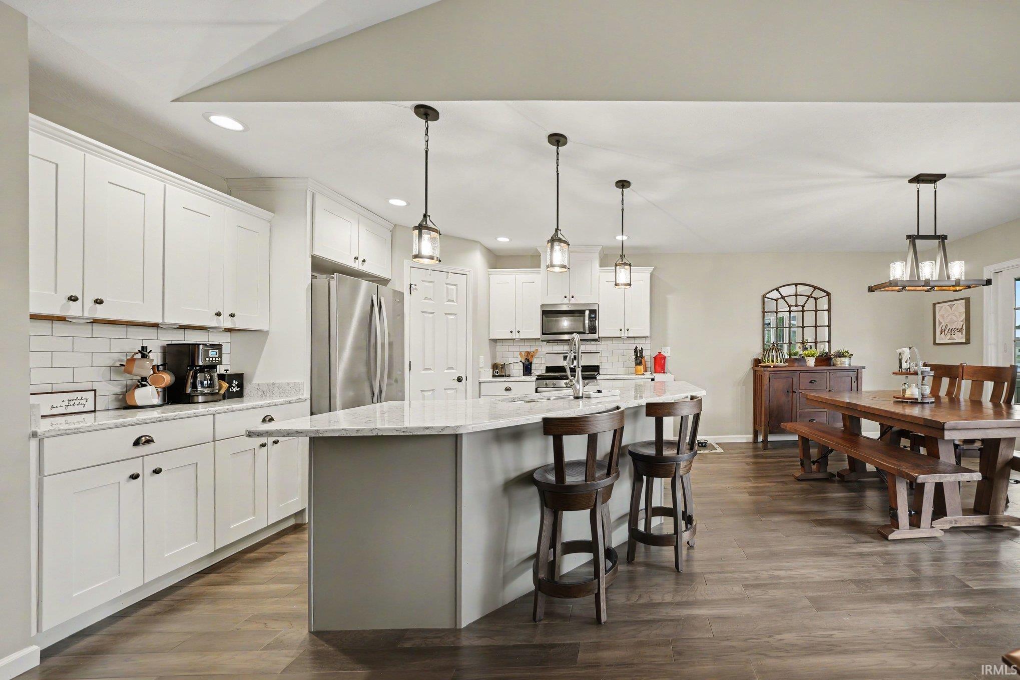 Expansive open-concept kitchen and dining area featuring wood-finish flooring, white shaker-style cabinetry, stainless steel appliances, a subway tile backsplash, and a large central island with seating