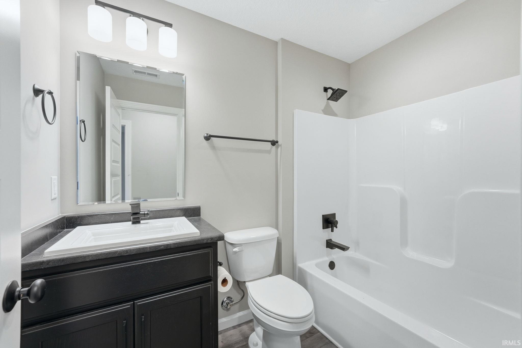 Bathroom featuring a dark wood-finish vanity with a white integrated sink, a rectangular mirror, a three-light vanity fixture, a toilet, and a tub/shower combination with matte black fixtures