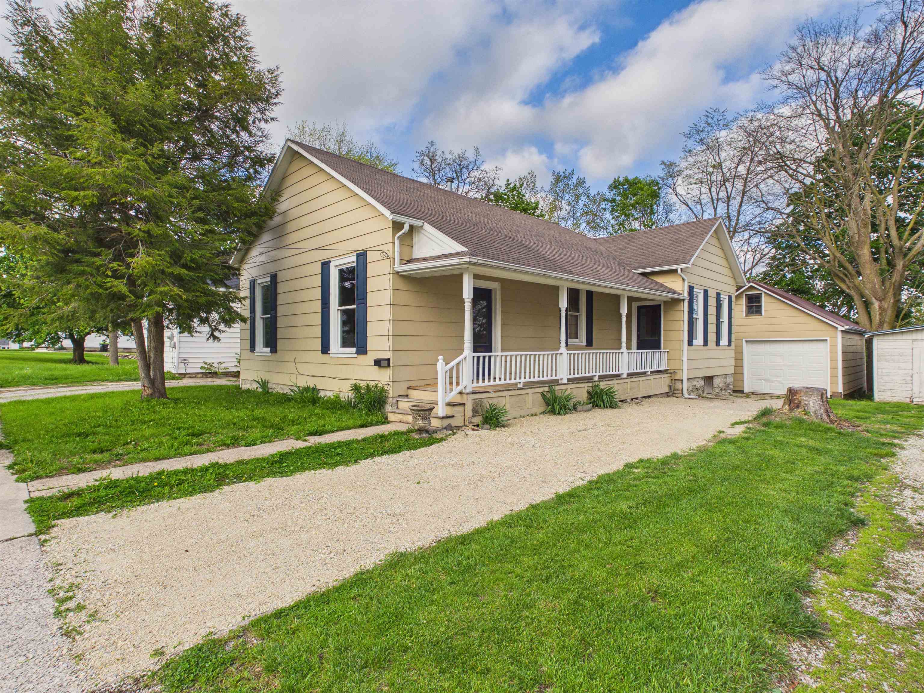 Single-story residence featuring light yellow siding, a covered front porch with white railing, blue window shutters, a gravel driveway, and an detached garage with a white overhead door