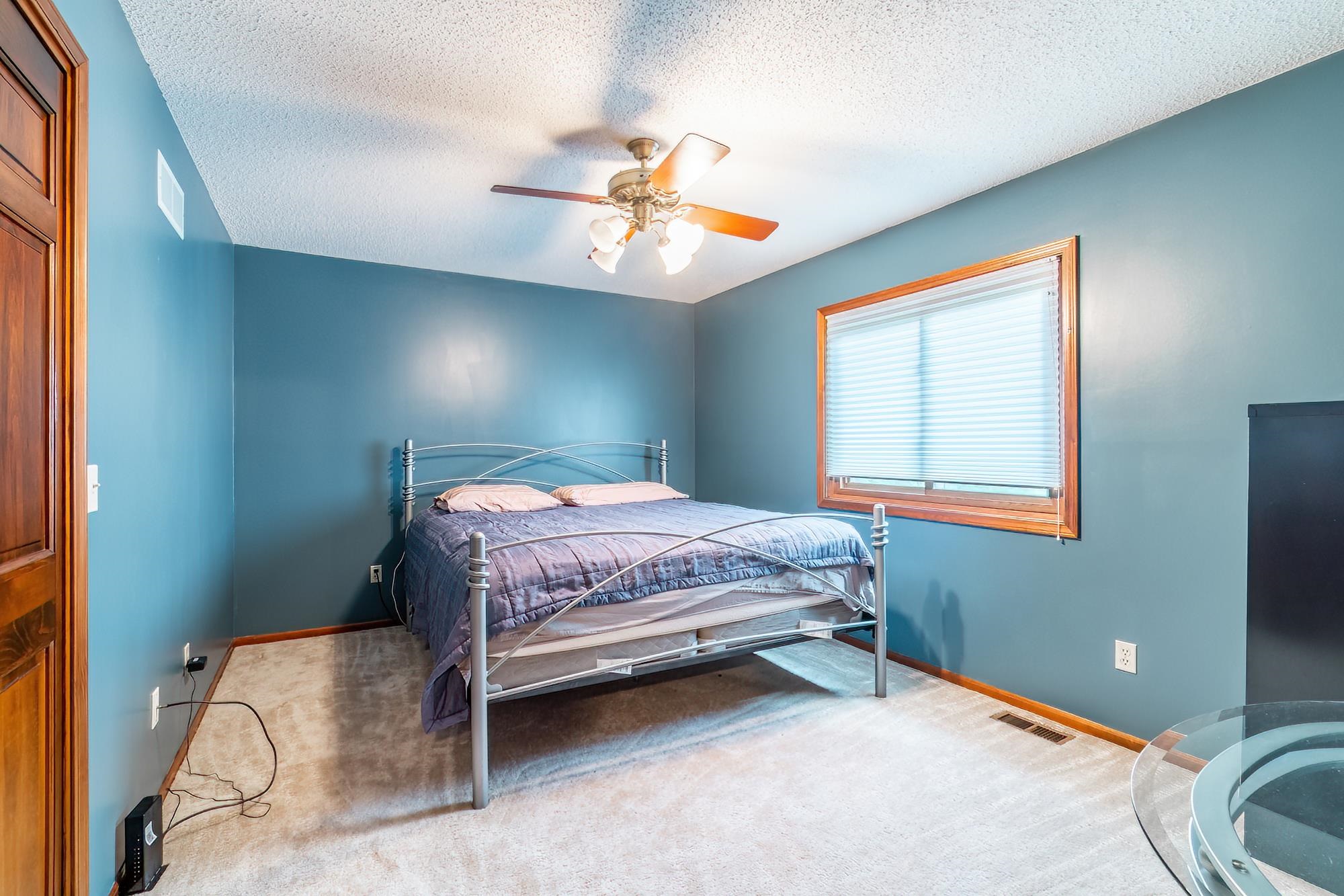 Bedroom featuring wall-to-wall carpeting, a ceiling fan with integrated lighting, wood-trimmed window with blinds, and a solid wood paneled door