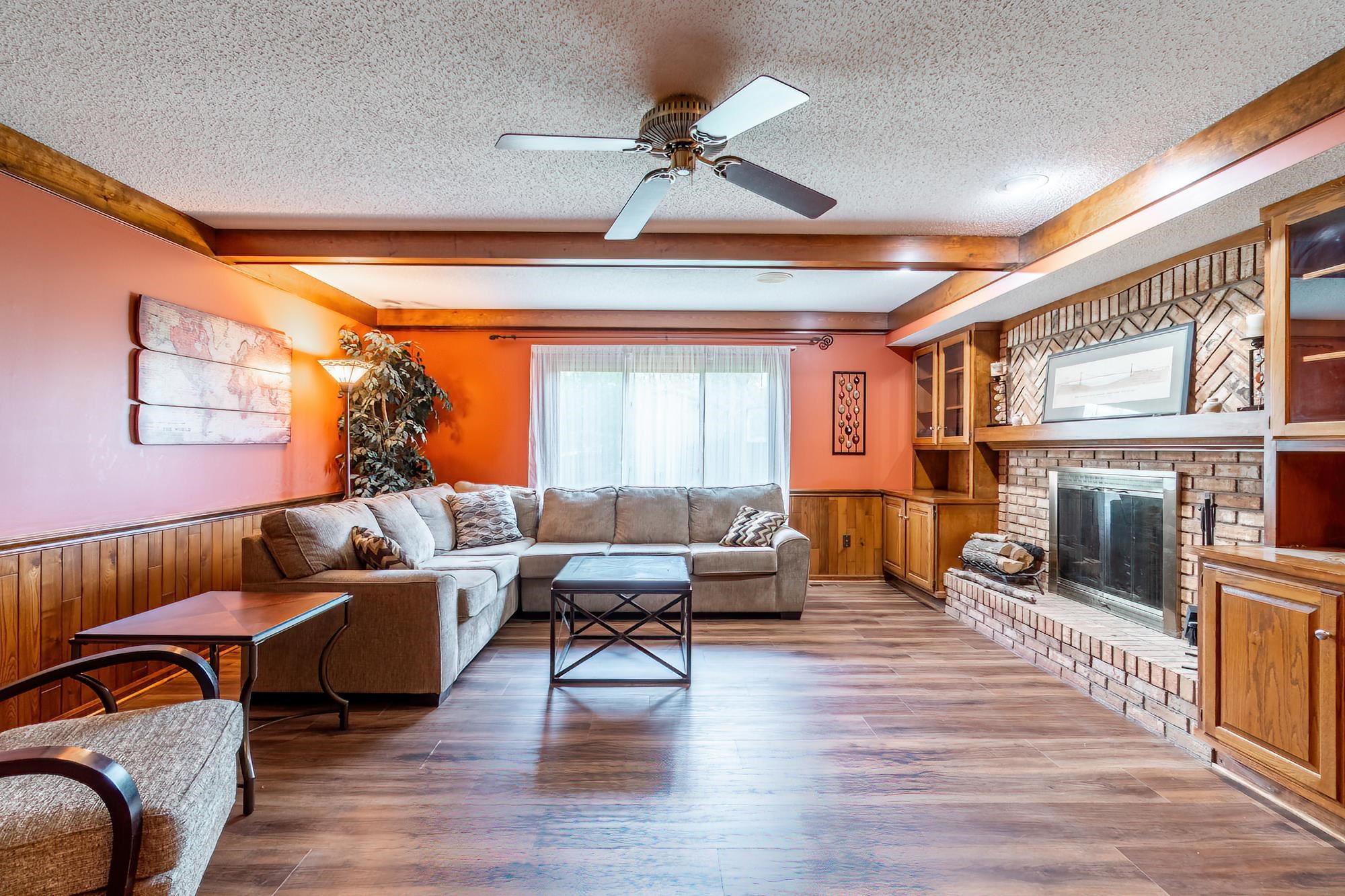 Living area featuring wood-finish flooring, a brick fireplace with a mantle, built-in cabinetry, wood beam accents, and a ceiling fan