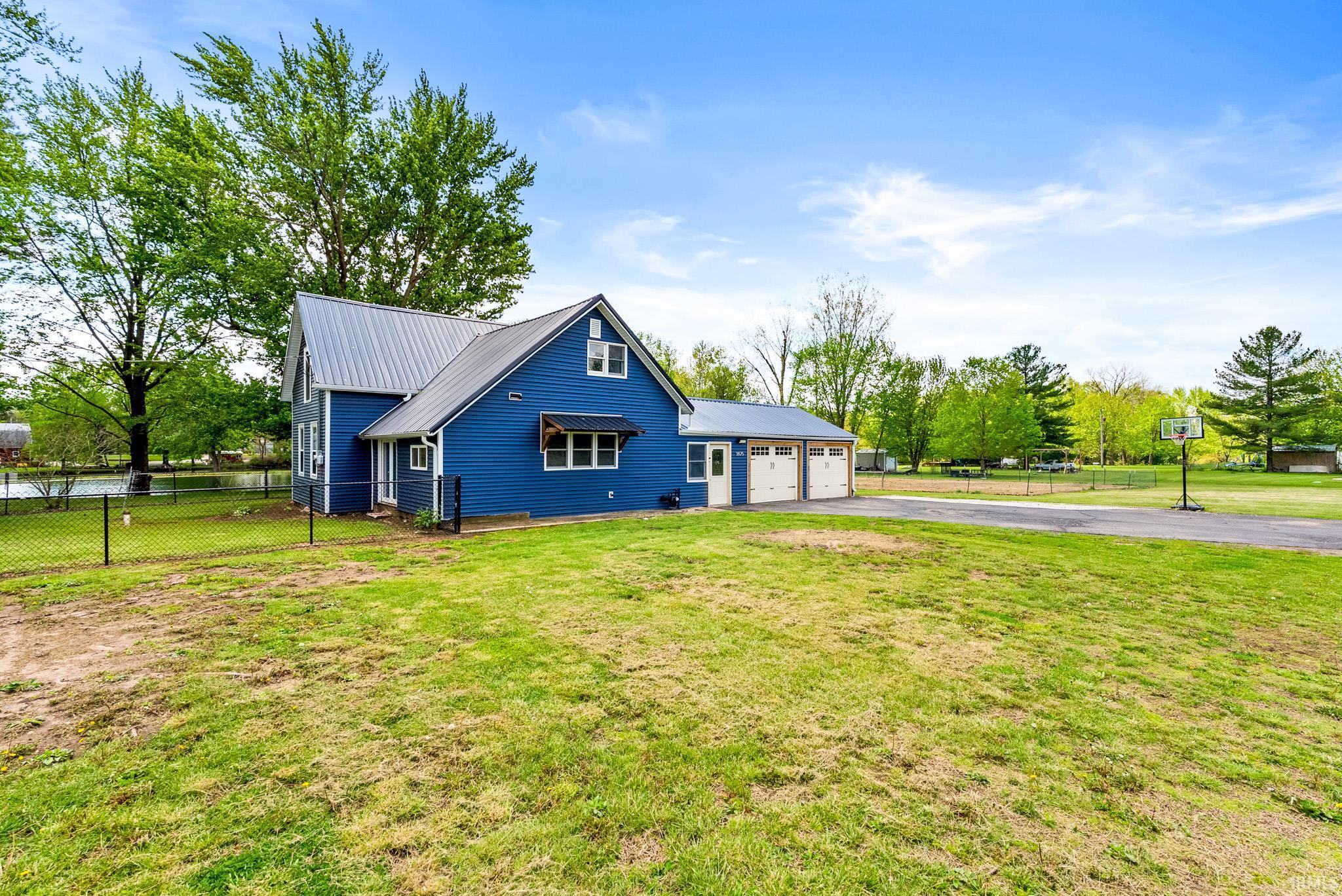 Blue siding exterior with a metal roof, featuring a two-car garage with white doors, a paved driveway, and a fenced yard