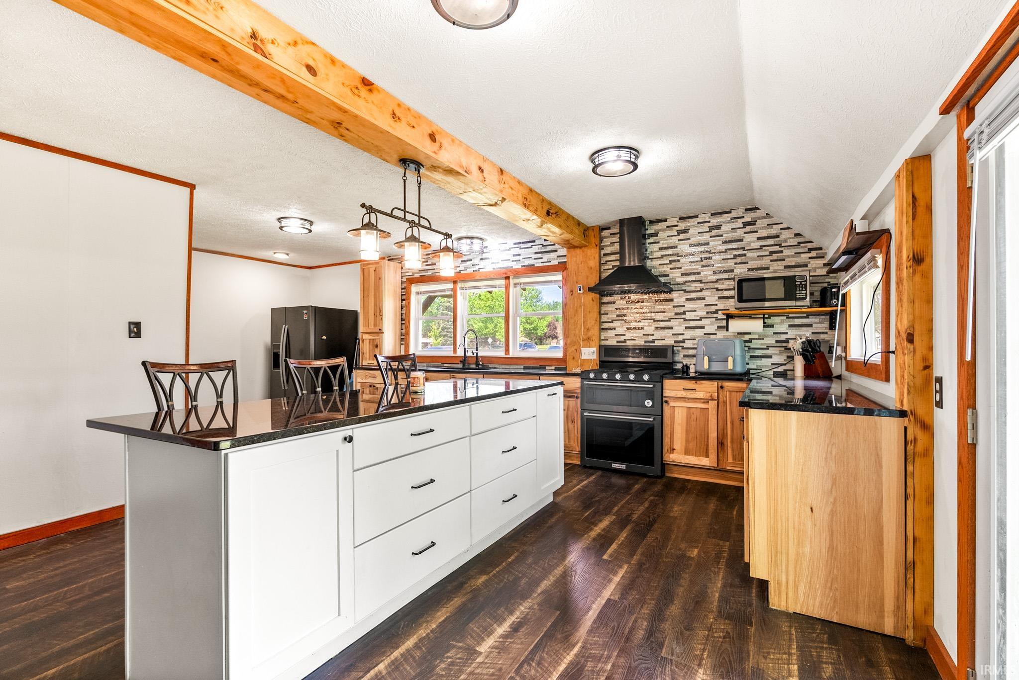Spacious kitchen featuring an extended island with white cabinetry and dark countertops, wood-finish flooring, exposed wood beams, and a tile backsplash