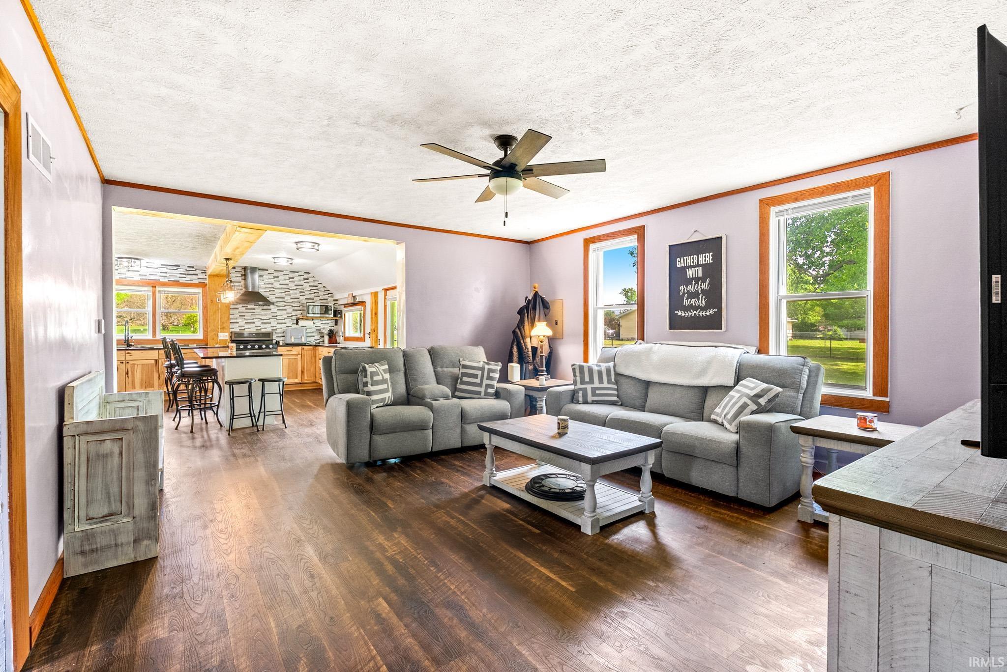 Open-concept living space featuring wood-finish flooring, a ceiling fan with integrated lighting, and multiple windows with wood trim