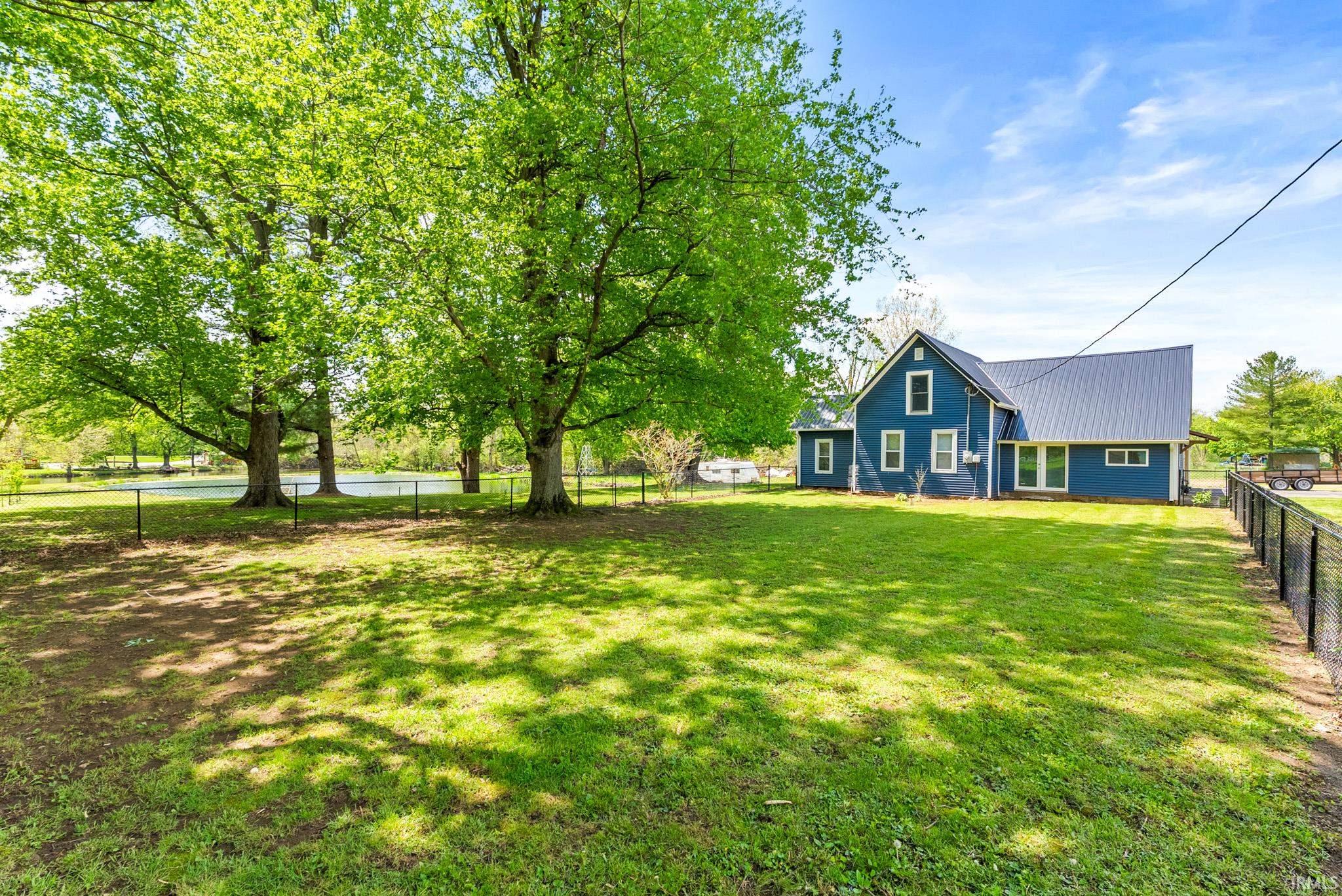 Expansive green lawn with mature shade trees, a perimeter fence, and a blue-sided residence featuring a dark metal roof and multiple windows