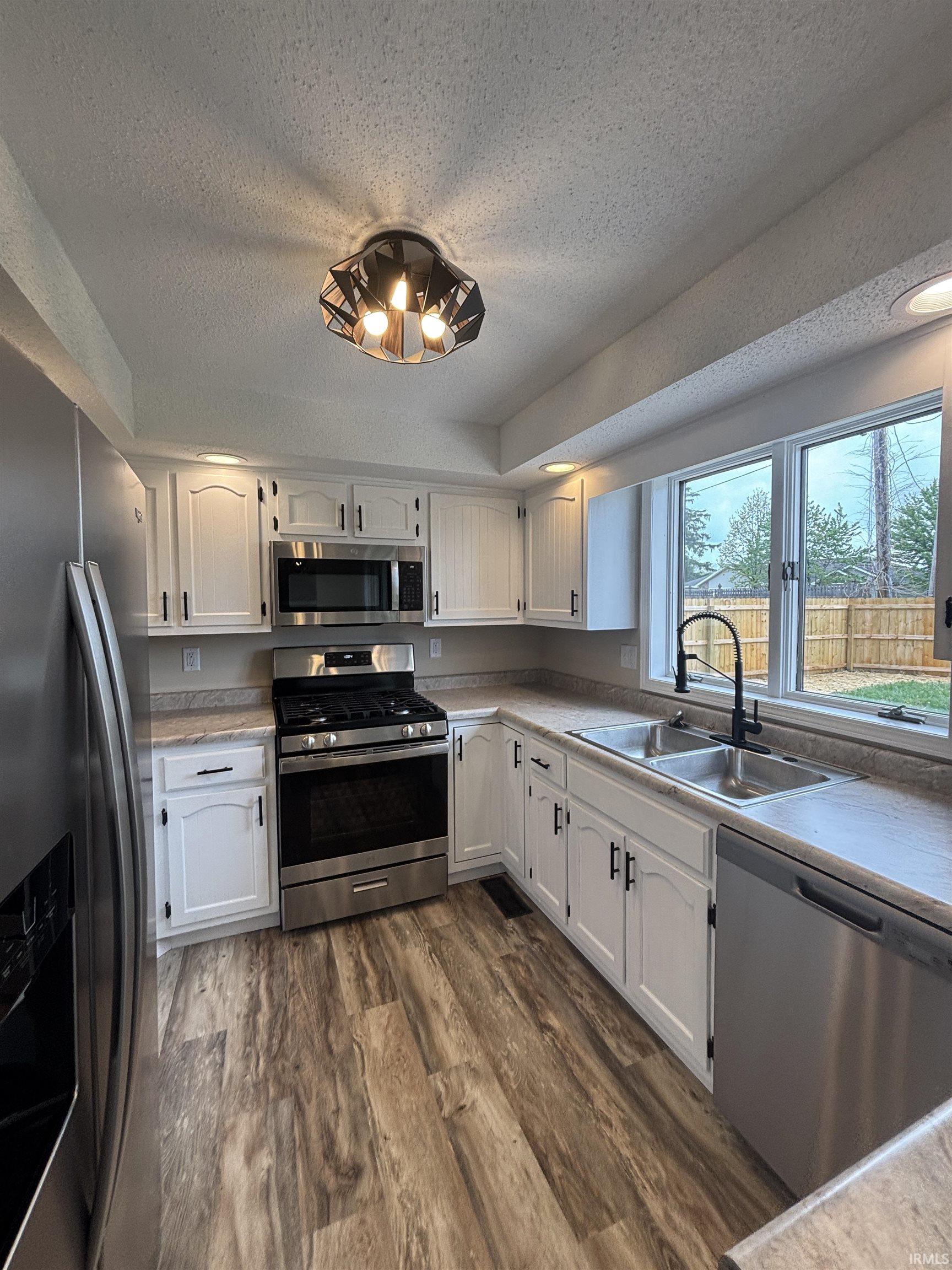 Modern kitchen featuring stainless steel appliances, white cabinetry with dark hardware, wood-finish flooring, a double basin sink with a gooseneck faucet, and ample natural light from a large window