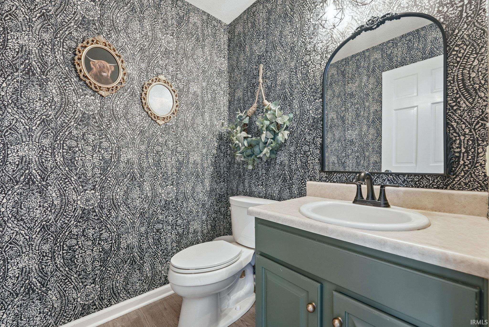 Powder room featuring patterned wallpaper, a dark green vanity with a light countertop, an oval undermount sink, and wood-finish flooring