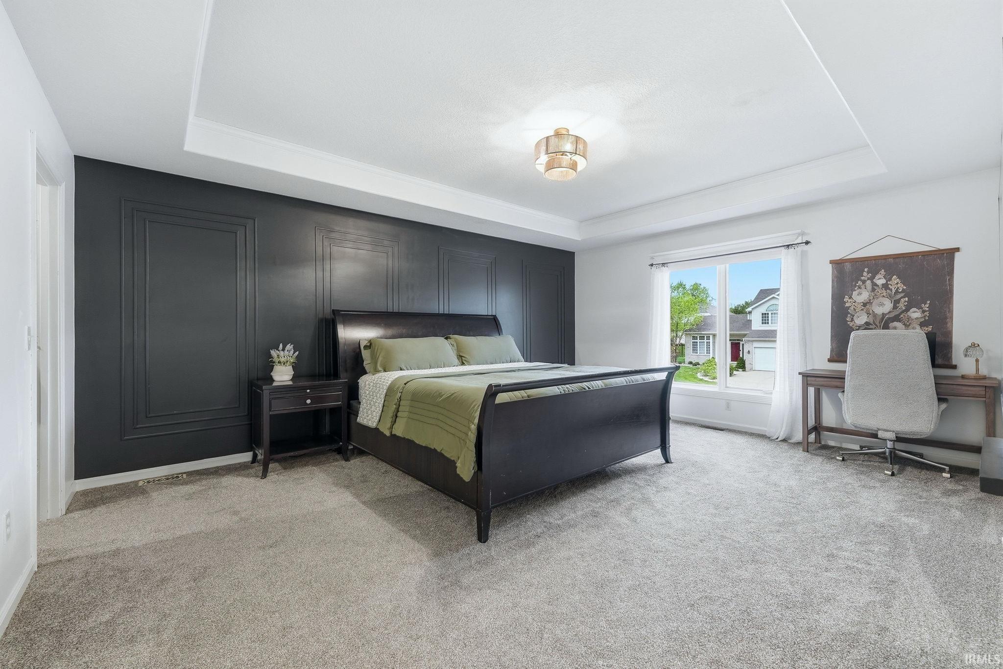 Spacious bedroom featuring a tray ceiling with a central light fixture, a paneled accent wall, a window with white trim, and plush carpet flooring