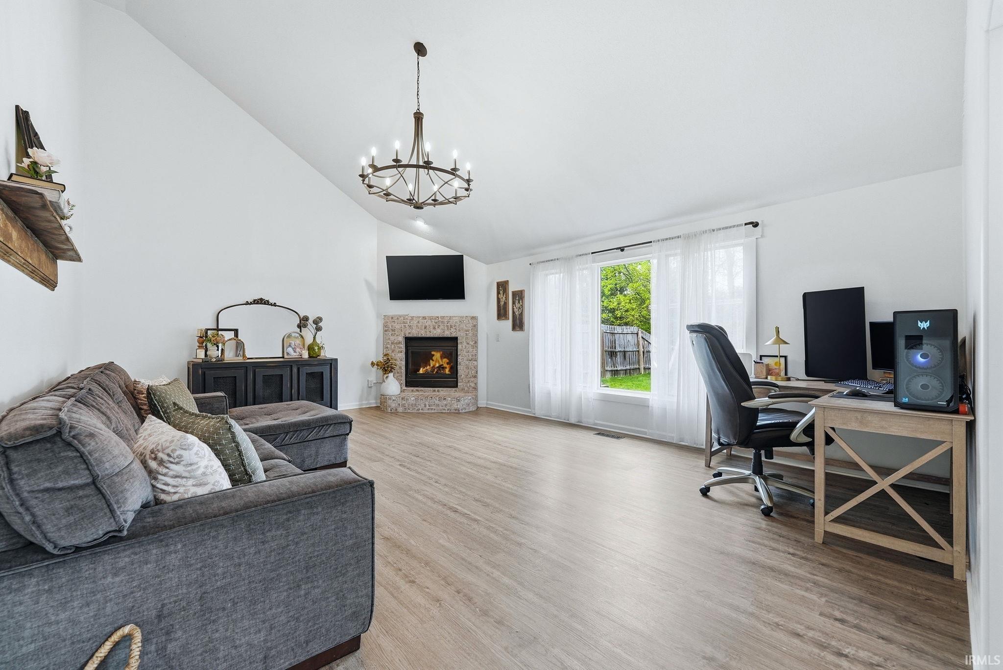 Spacious living area featuring a vaulted ceiling with an iron chandelier, light wood-finish flooring, and a corner fireplace with brick surround