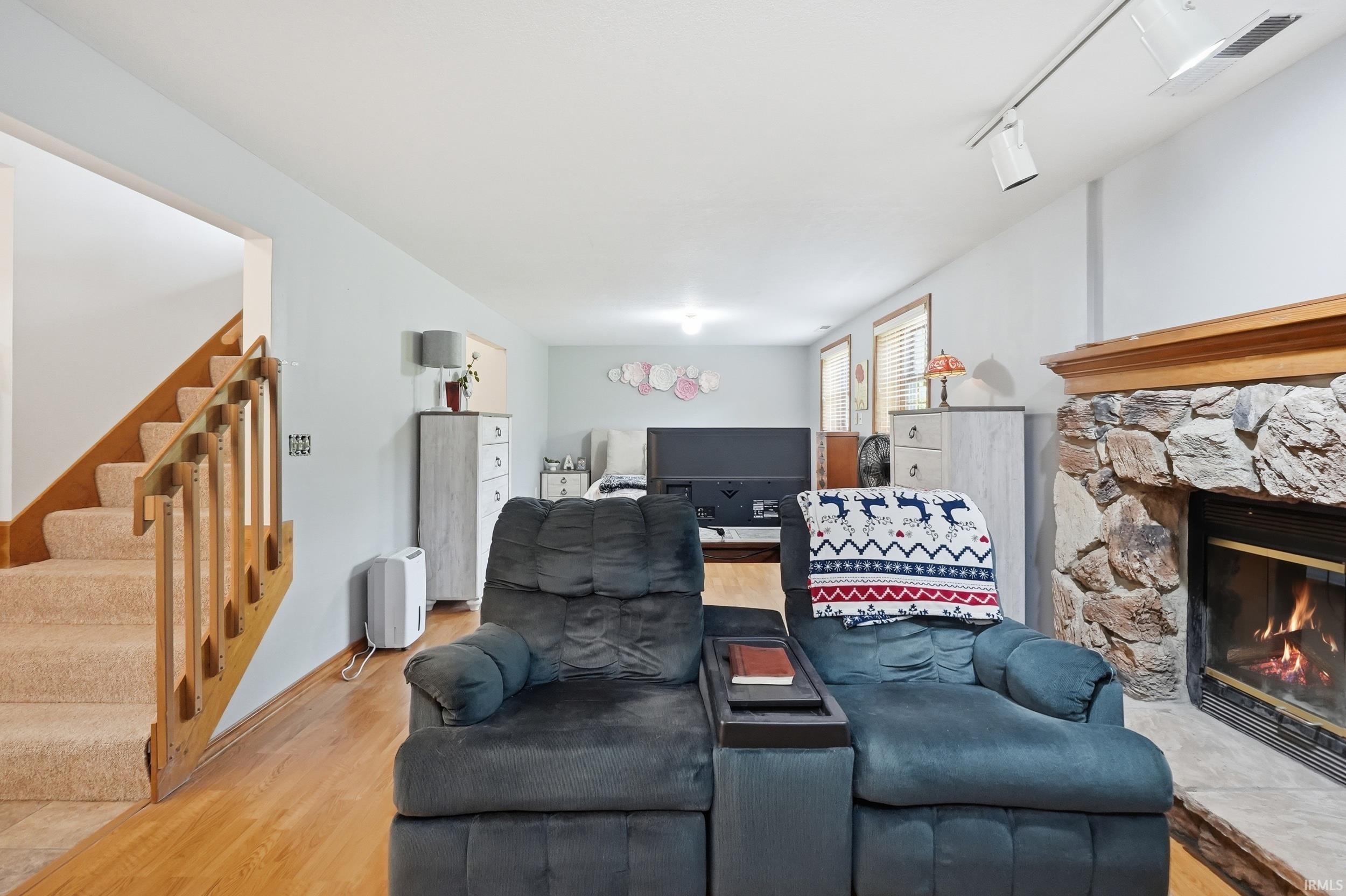 Stone fireplace with a wooden mantel, wood-finish flooring, staircase with carpeted treads and wood railings, track lighting, and recessed lighting