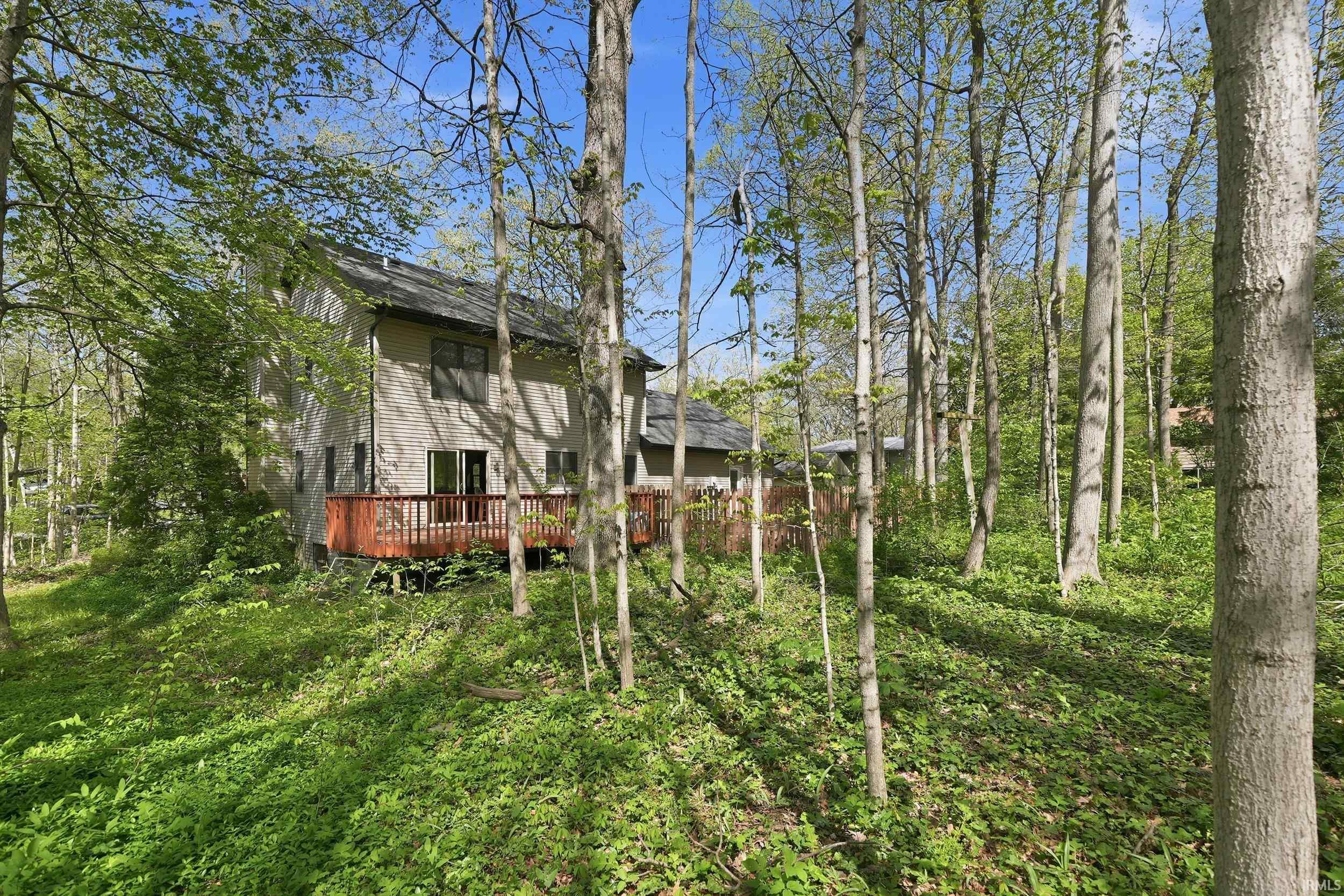 Two-story residence with a multi-level deck, dark shingle roof, and light-toned siding, set amidst dense deciduous trees and abundant ground cover