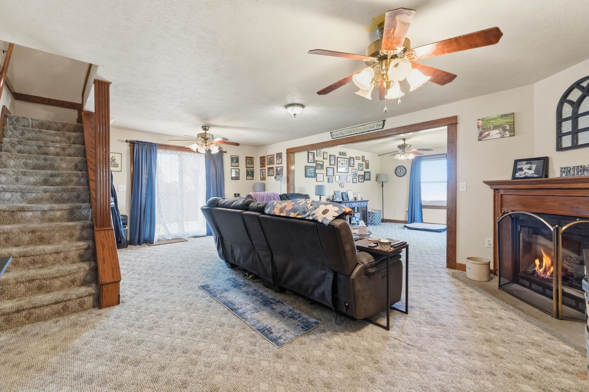 Spacious living area featuring a fireplace with a wood mantle, carpet flooring, a ceiling fan, and a sliding glass door