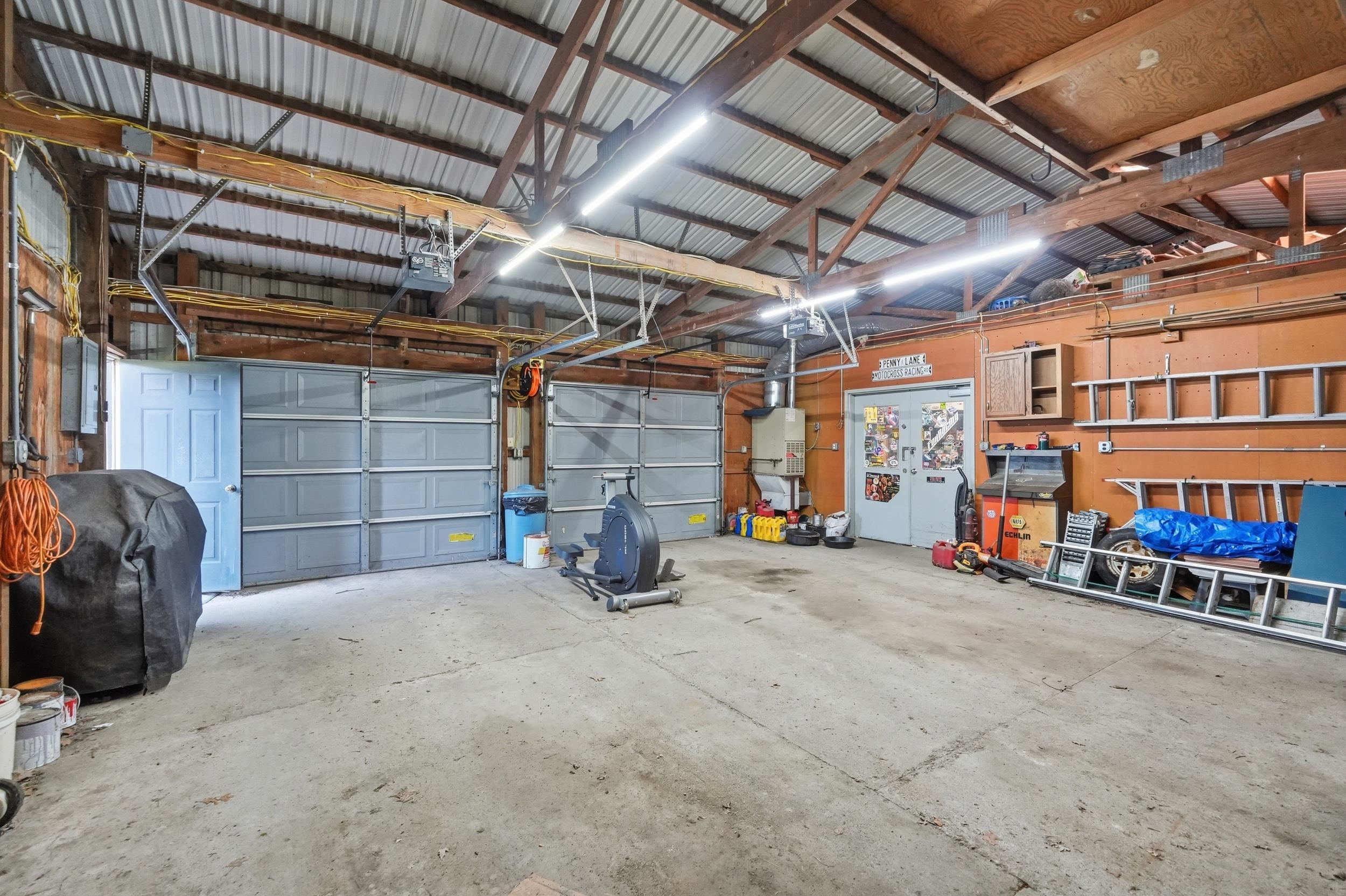 Spacious garage featuring two overhead doors, a pedestrian door, exposed wood rafters, corrugated metal roofing, and fluorescent lighting fixtures