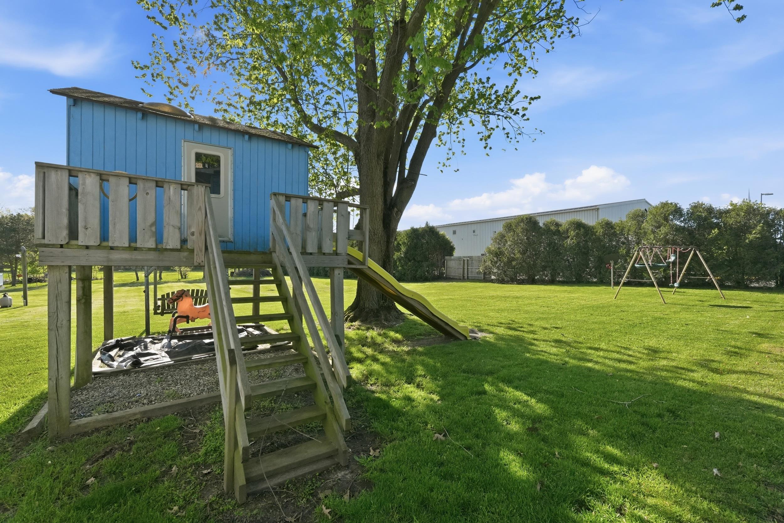 Expansive green lawn featuring a built-in playhouse with wooden stairs and deck, an attached slide, and a separate swing set