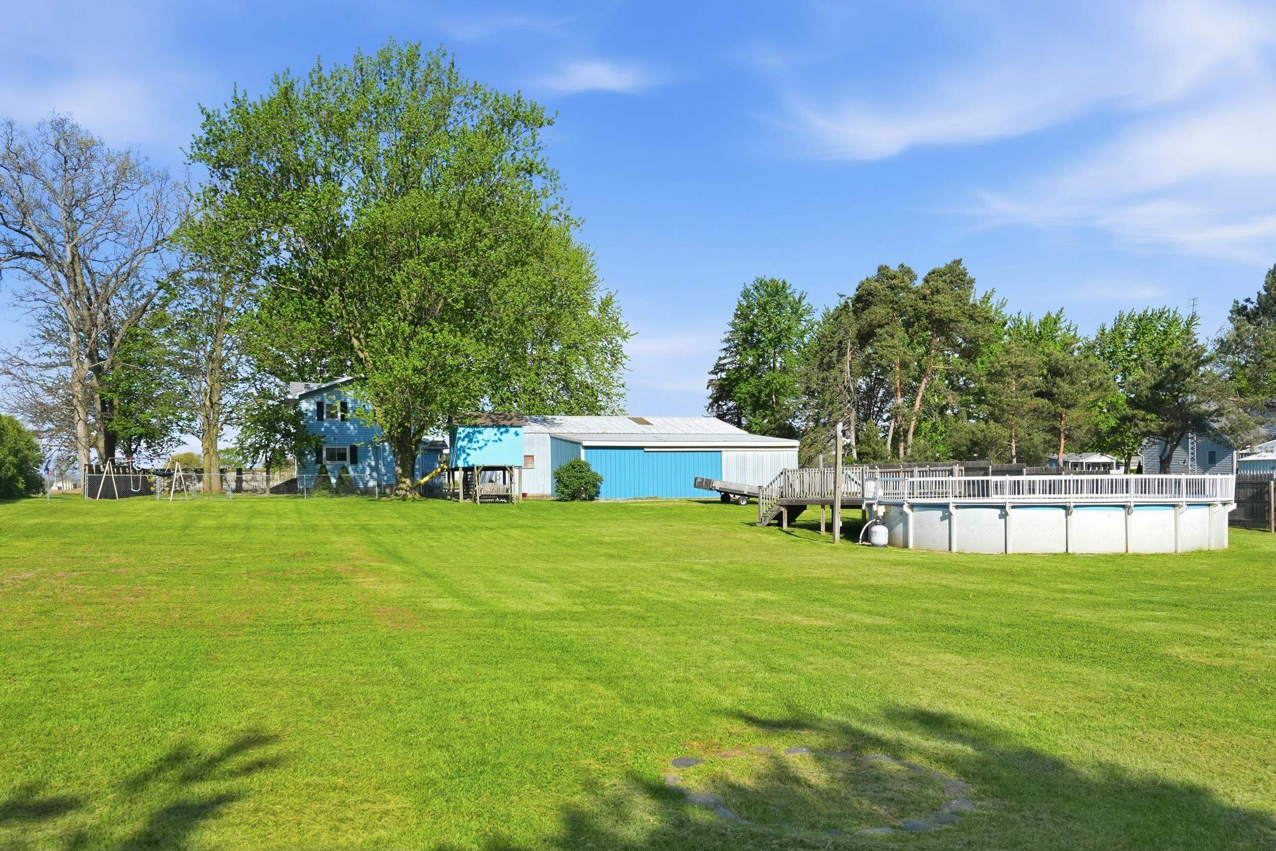 Expansive green lawn with mature trees, above-ground pool with surrounding wood decking, and a light blue structure with a bright blue outbuilding