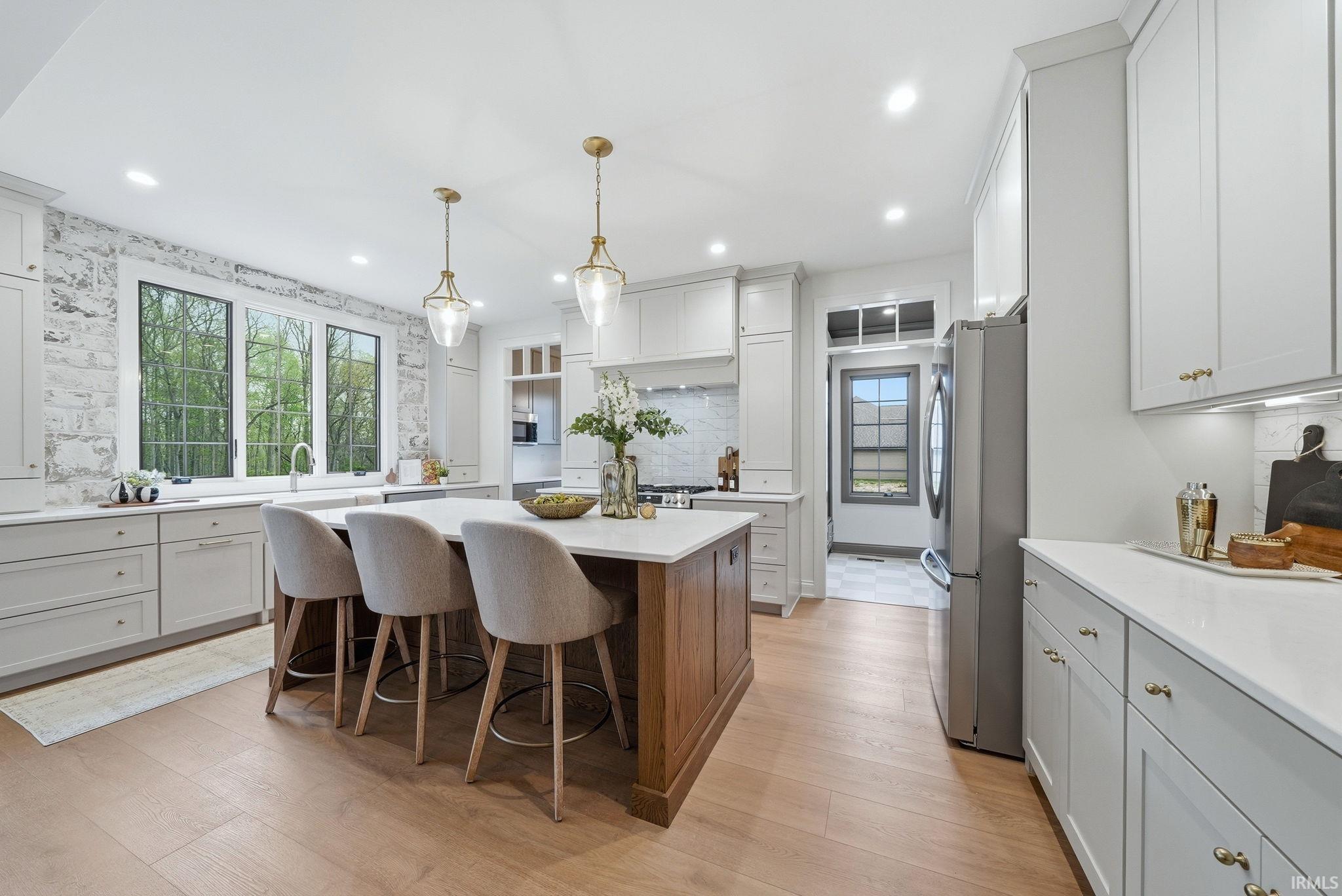 Contemporary kitchen featuring a substantial wood-finish island with seating, light wood-finish flooring, white cabinetry with gold-tone hardware, recessed lighting, and a white brick-finish accent wall