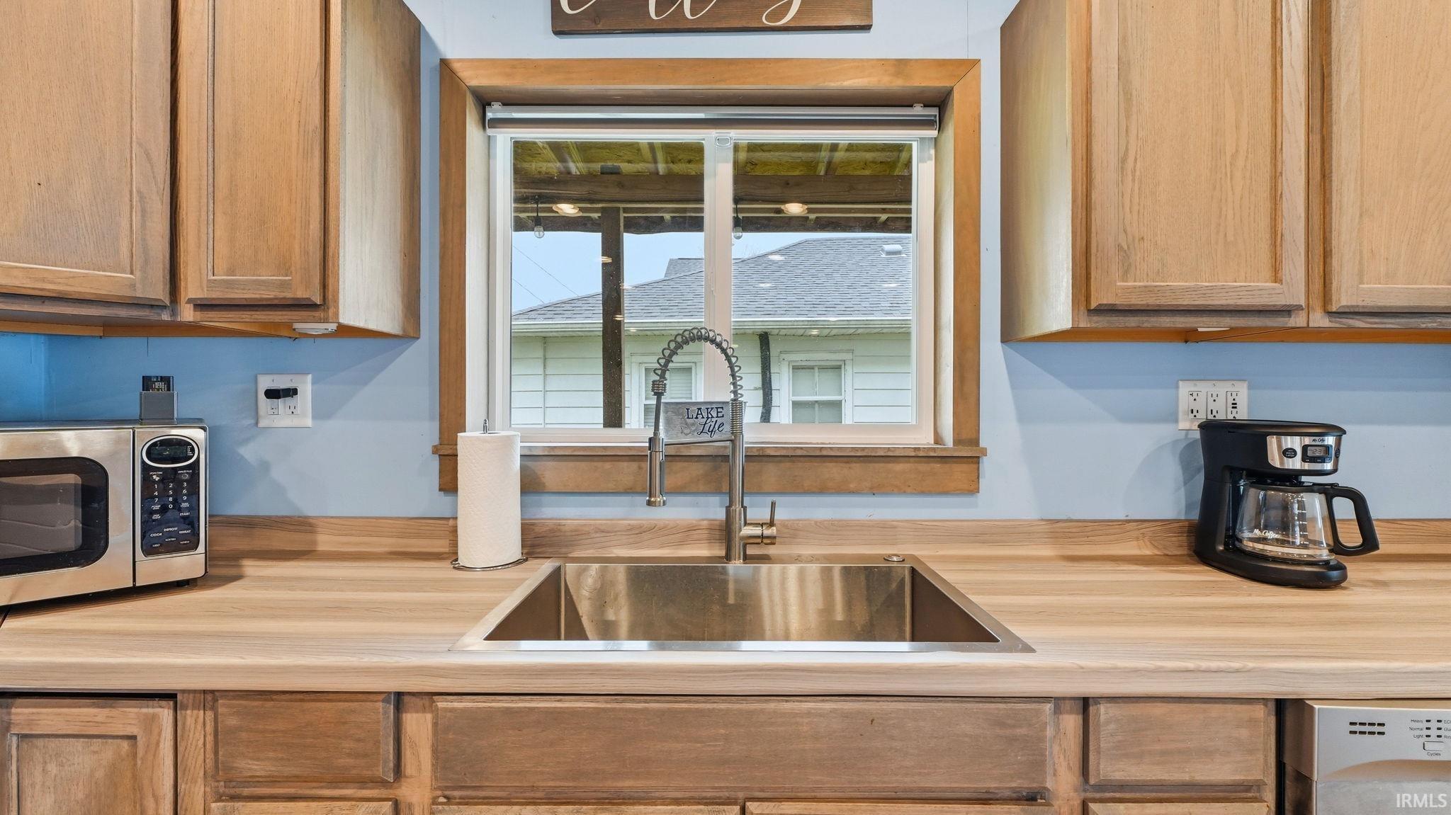 Kitchen sink area featuring a stainless steel undermount sink, gooseneck faucet, light wood-finish cabinetry, and a window with wood trim