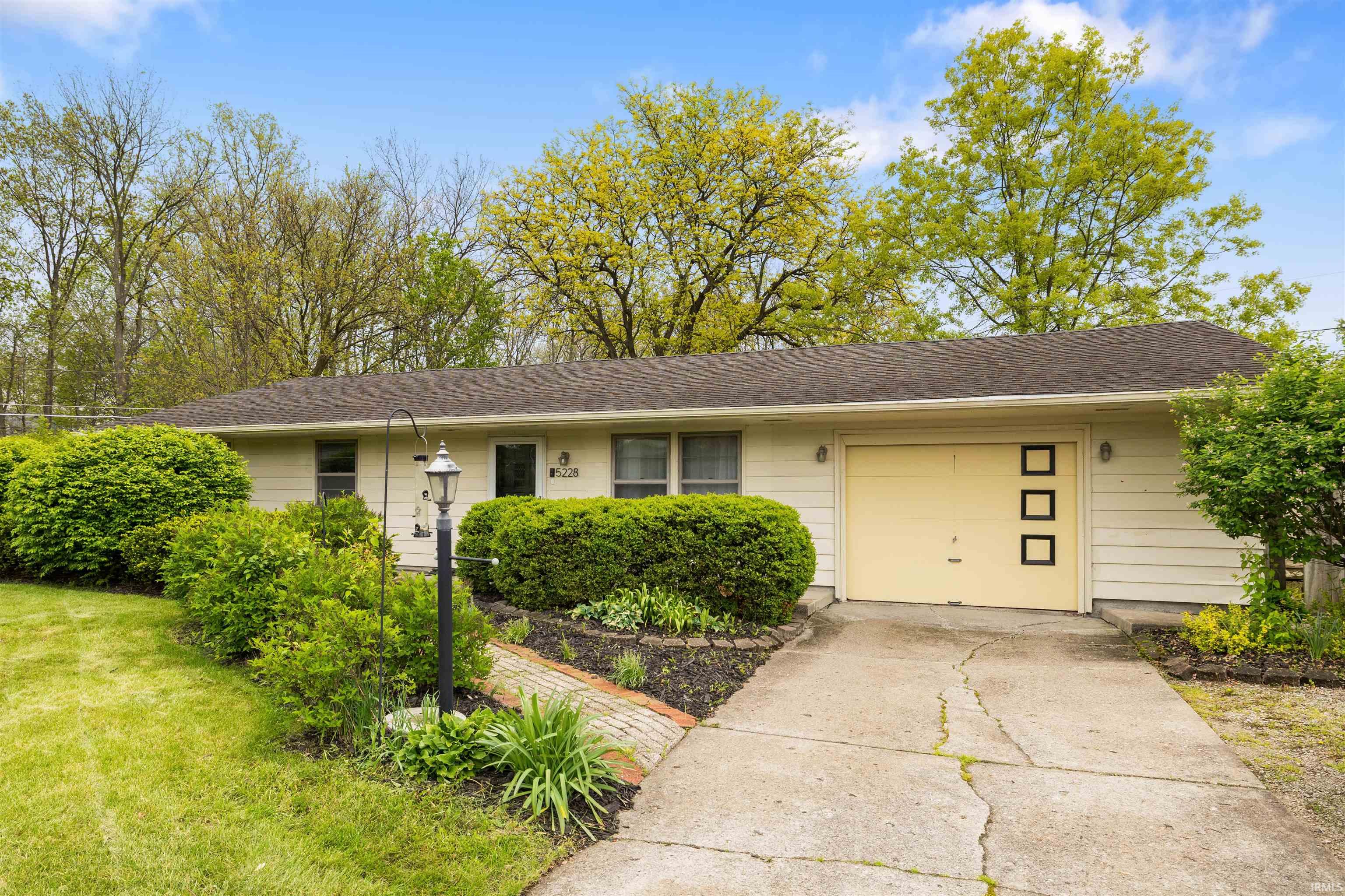 Ranch-style exterior with light-colored siding, an attached garage, mature landscaping, a paved driveway, and a brick-edged planting bed