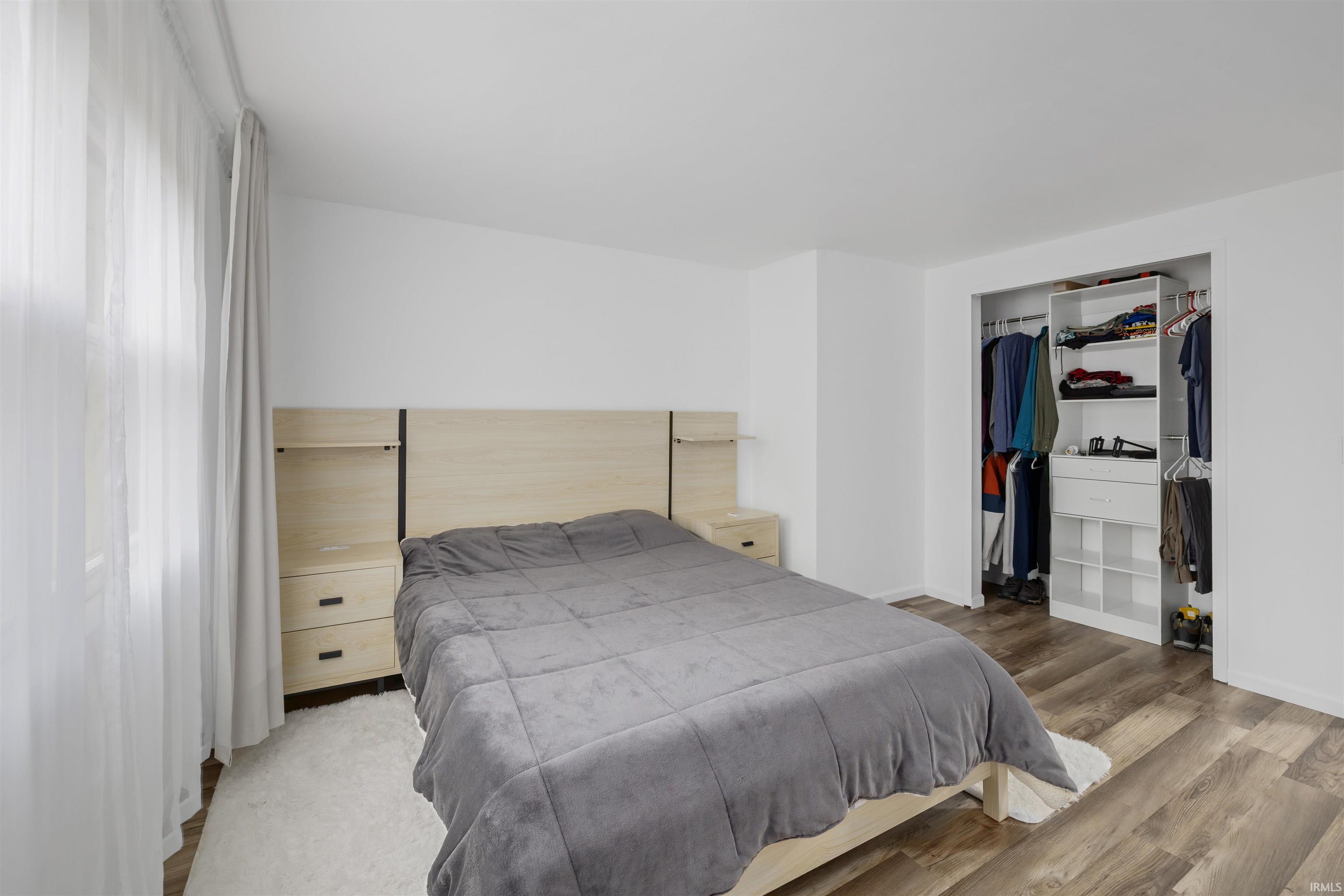 Bedroom featuring wood-finish flooring, a window with sheer and blackout curtains, and a built-in closet system with shelving and hanging rods
