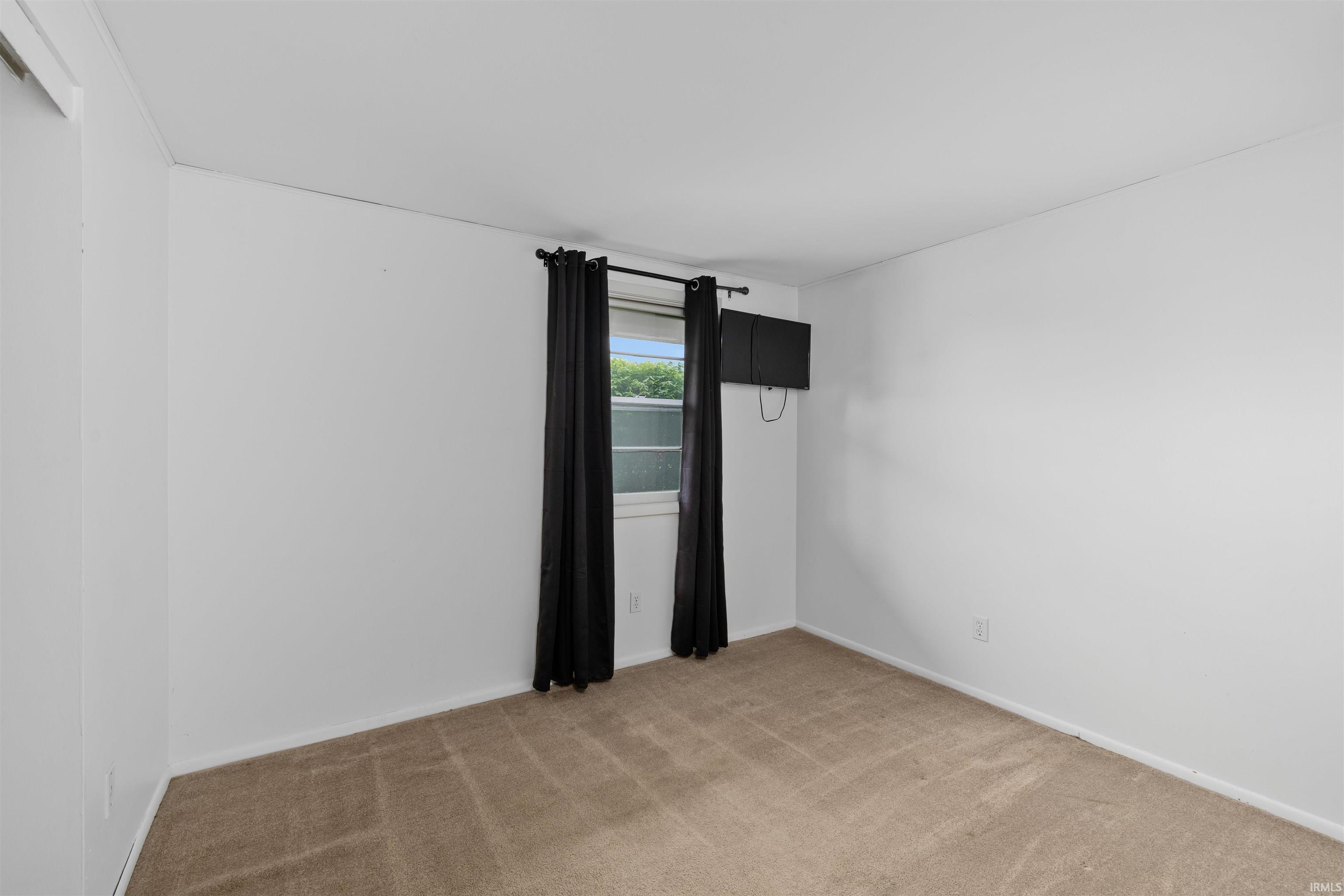 Carpeted interior room featuring white painted walls, a single window with black drapery, and a wall-mounted television