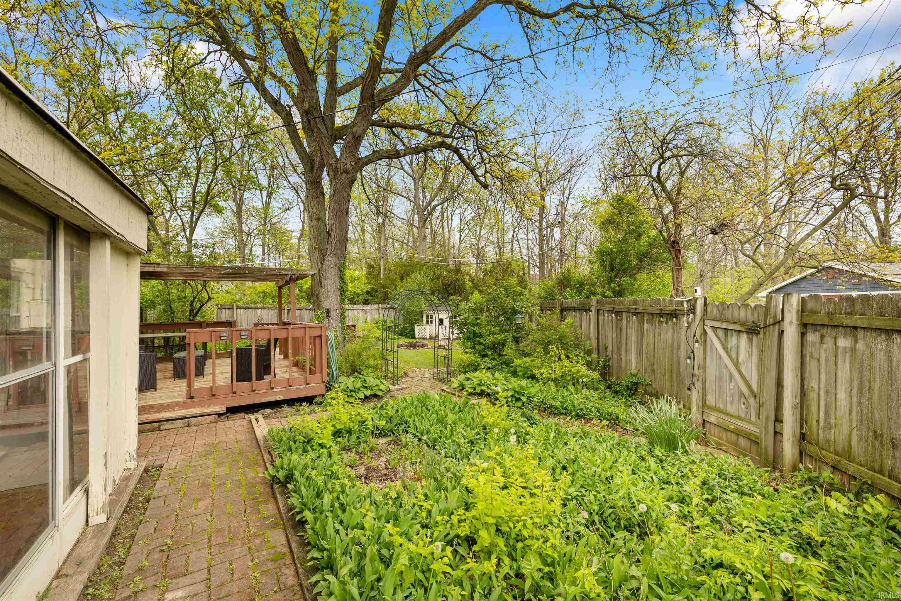 Expansive backyard featuring a wood deck with pergola, brick paver patio, a lush garden area, and mature trees providing natural shade