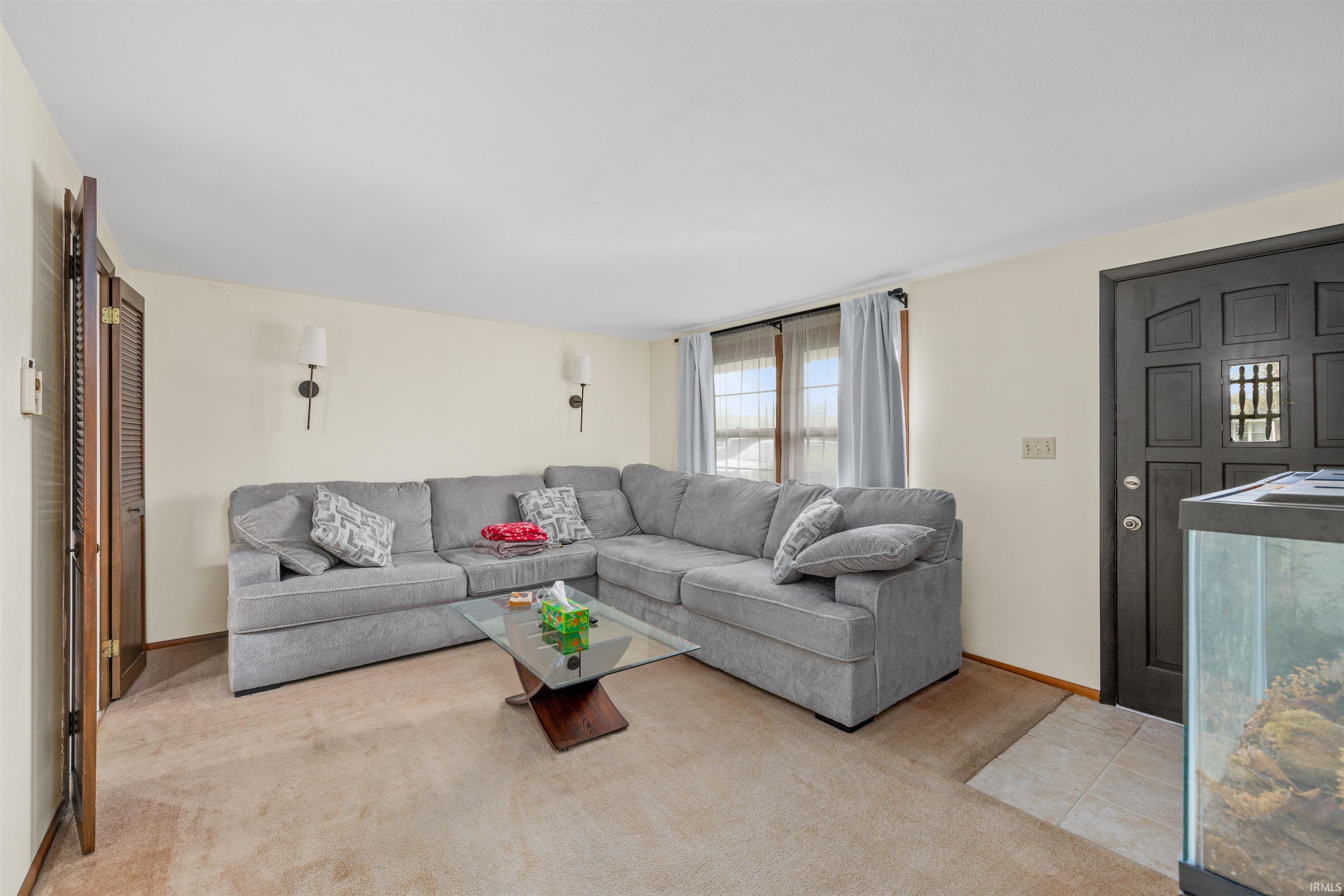 Carpeted living area featuring a front entry door with a decorative window, two wall sconces, and a window with curtain rods