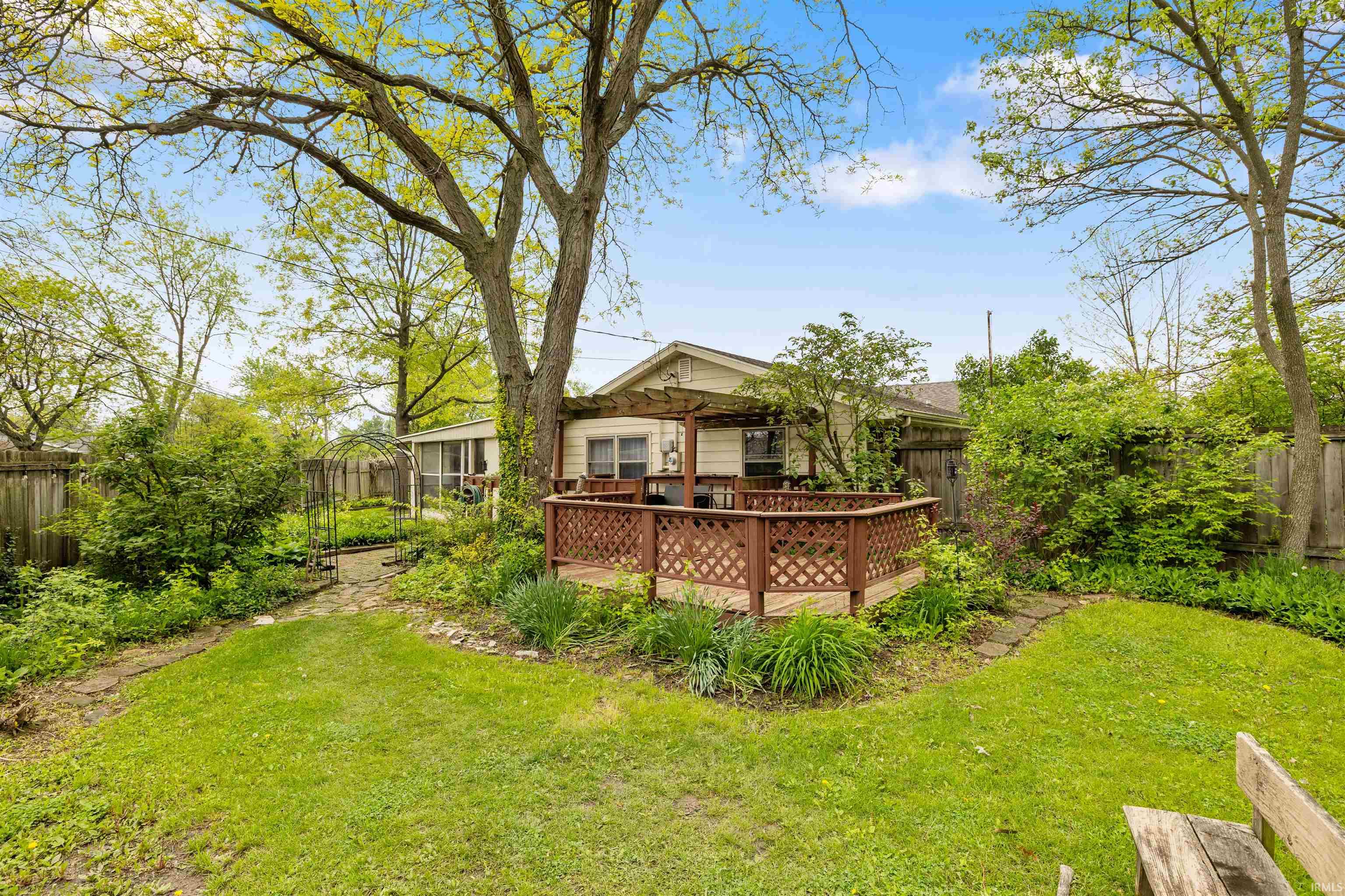 Rear exterior featuring a wooden deck with lattice railing and pergola, surrounded by mature trees and established greenery