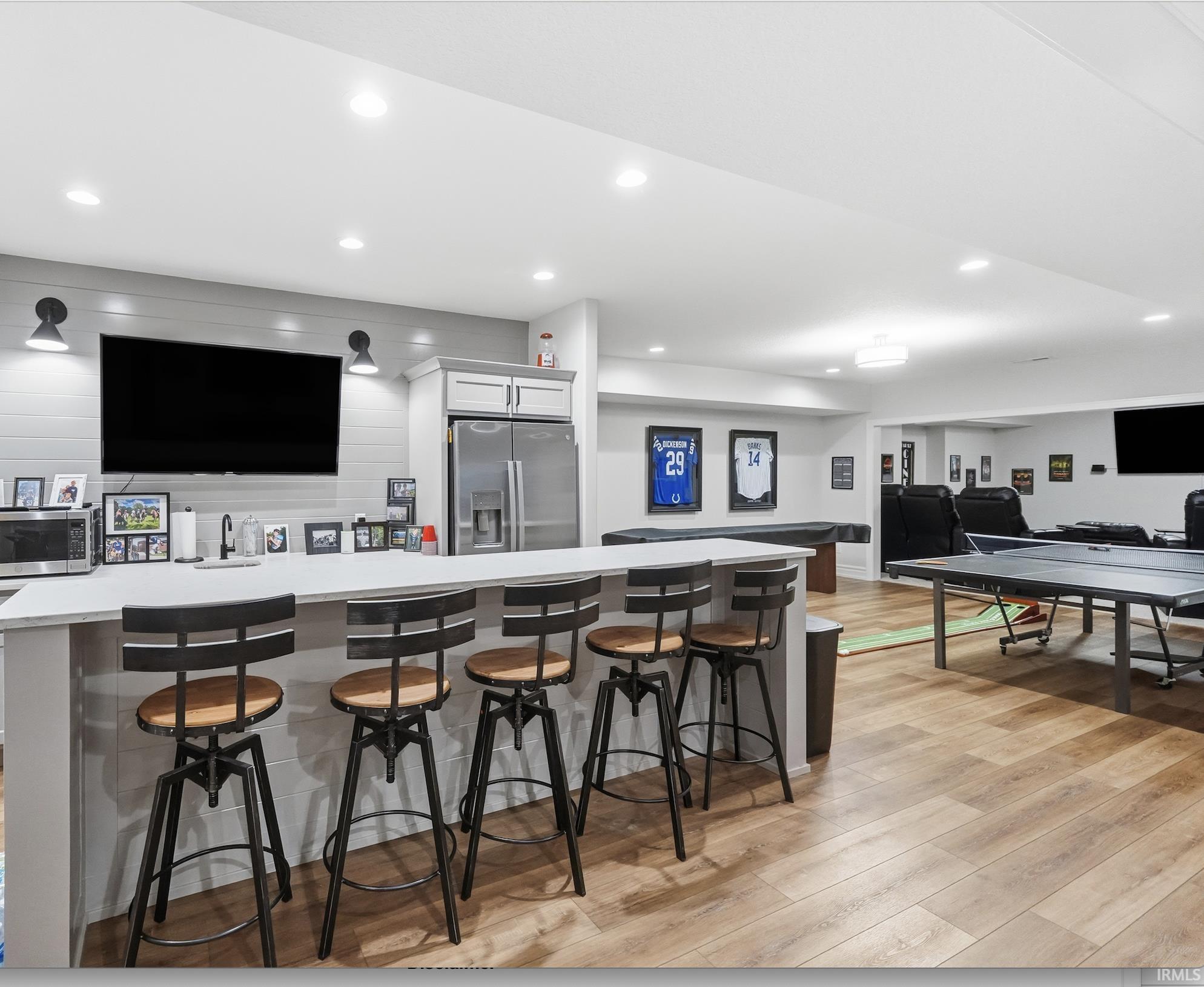 Spacious entertainment area featuring a wet bar with integrated stainless steel refrigerator, recessed lighting, wood-finish flooring, and shiplap accent wall