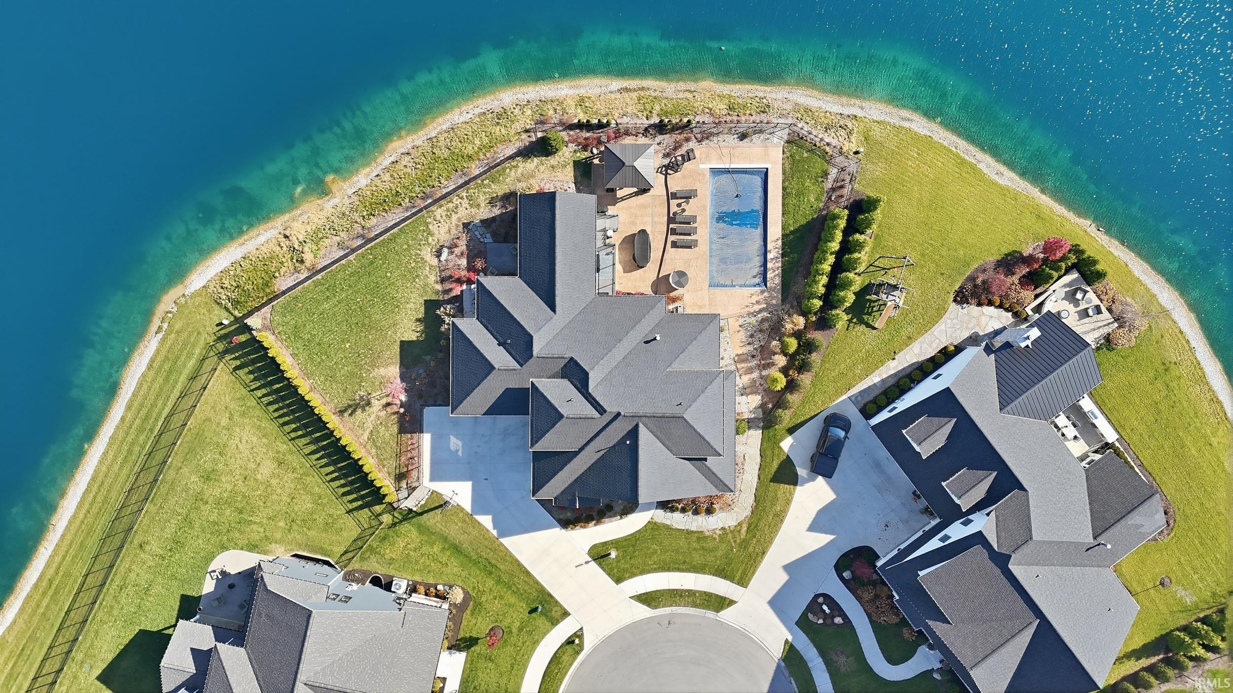 Expansive waterfront property featuring a large in-ground swimming pool, a spacious patio area with an outdoor kitchen, lush green lawns, and a circular driveway