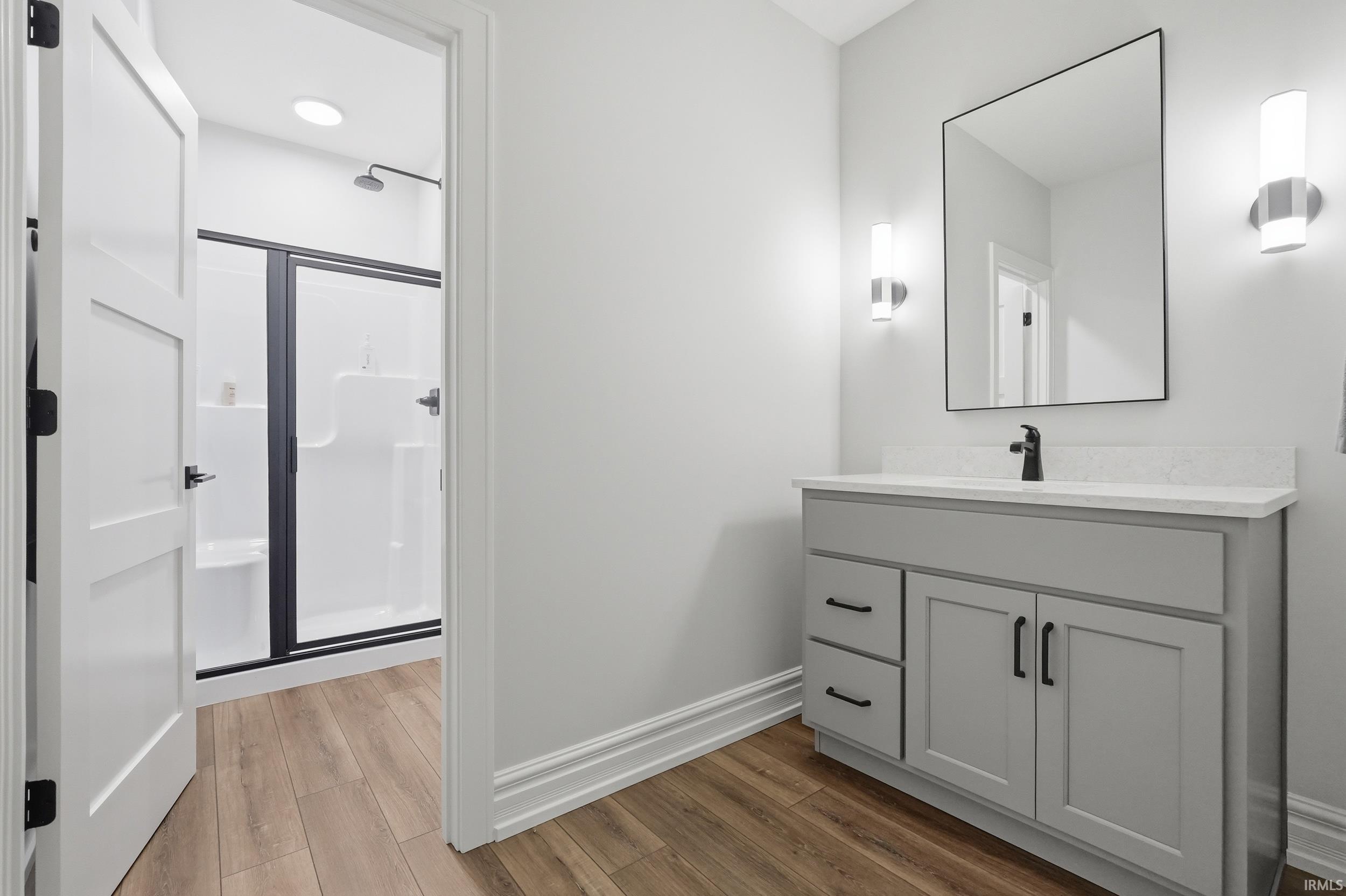 Bathroom featuring a gray vanity with a white countertop, a rectangular framed mirror, matte black fixtures, wood-finish flooring, and a shower with black-framed glass doors