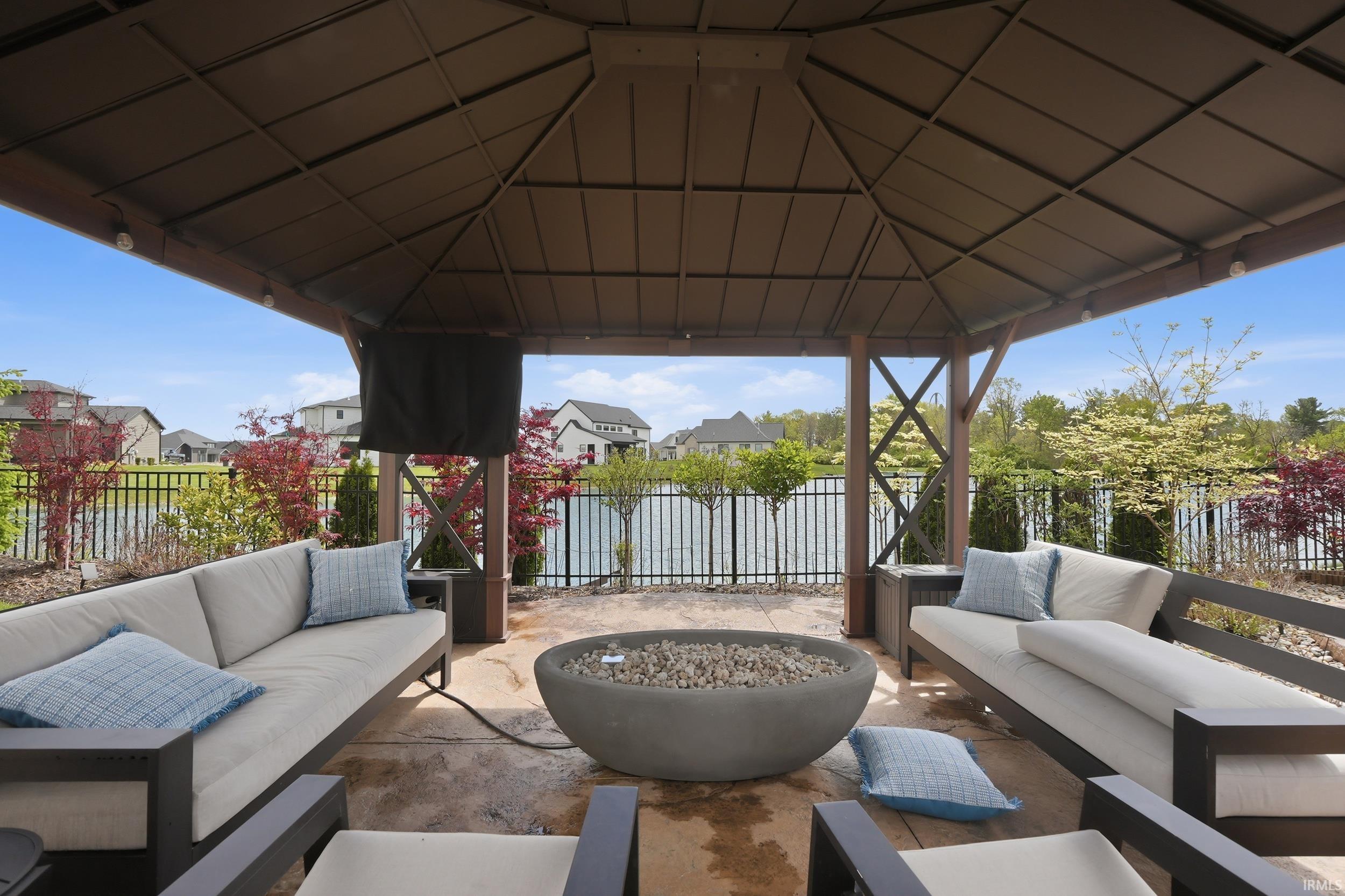 Covered outdoor patio featuring a central fire pit, concrete flooring, and a black metal fence bordering a water feature