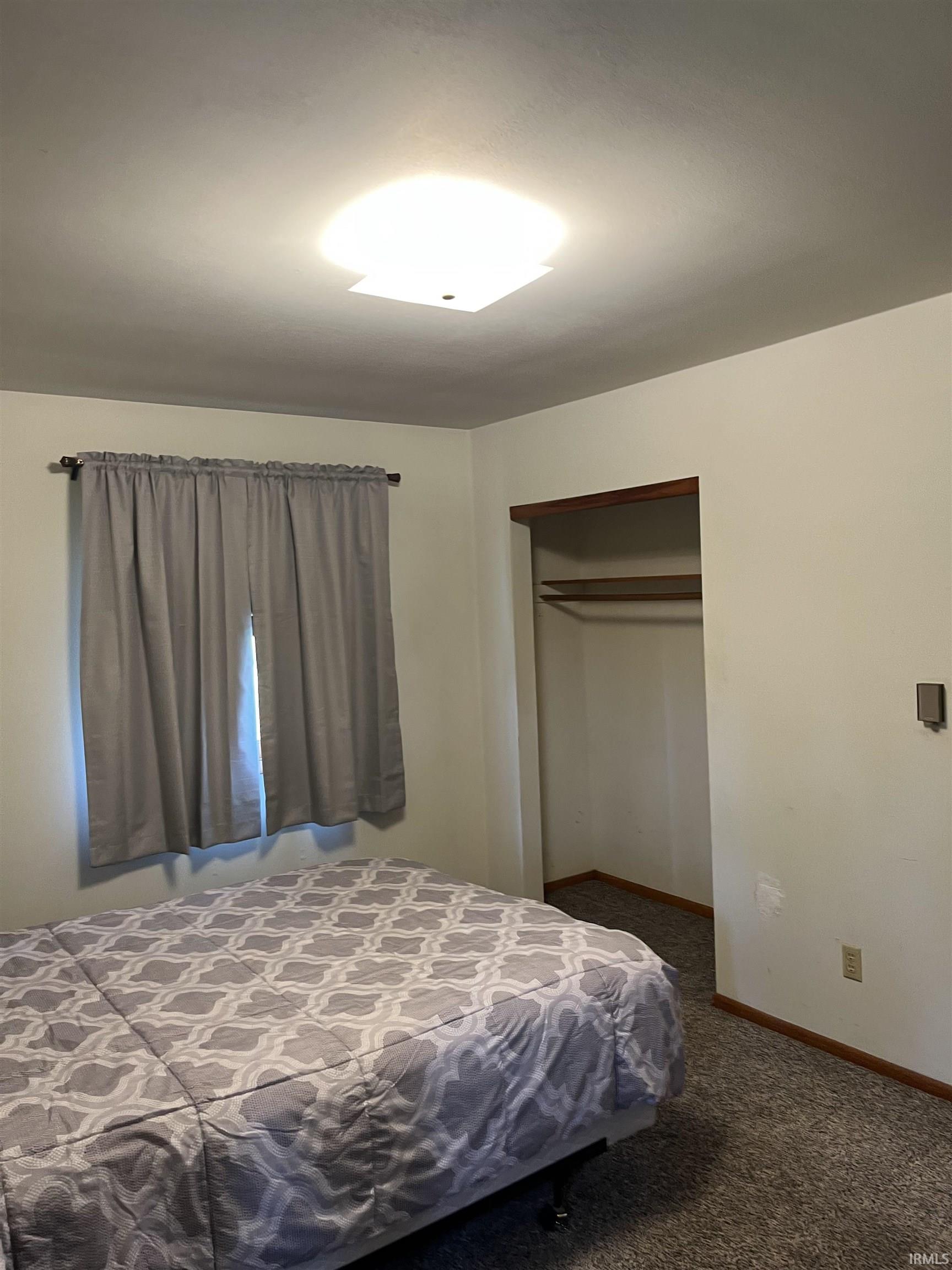 Carpeted room with a square ceiling light fixture, a window with curtains, and an open closet featuring a hanging rod and upper shelf