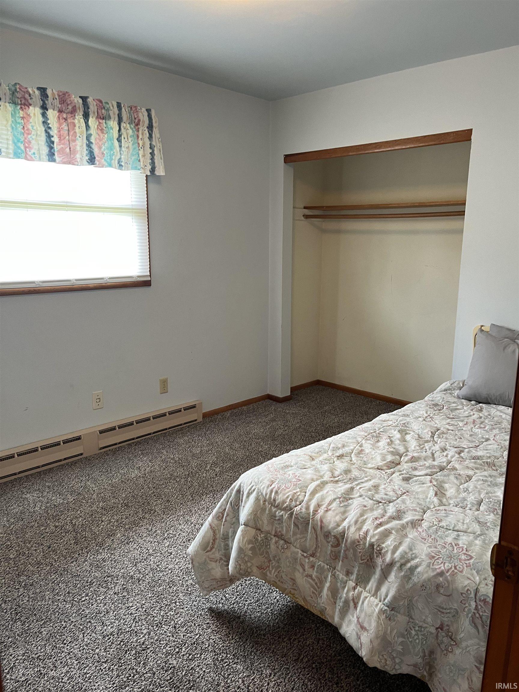 Carpeted room featuring a window with blinds, a baseboard heater, and an open closet with shelving and hanging rods