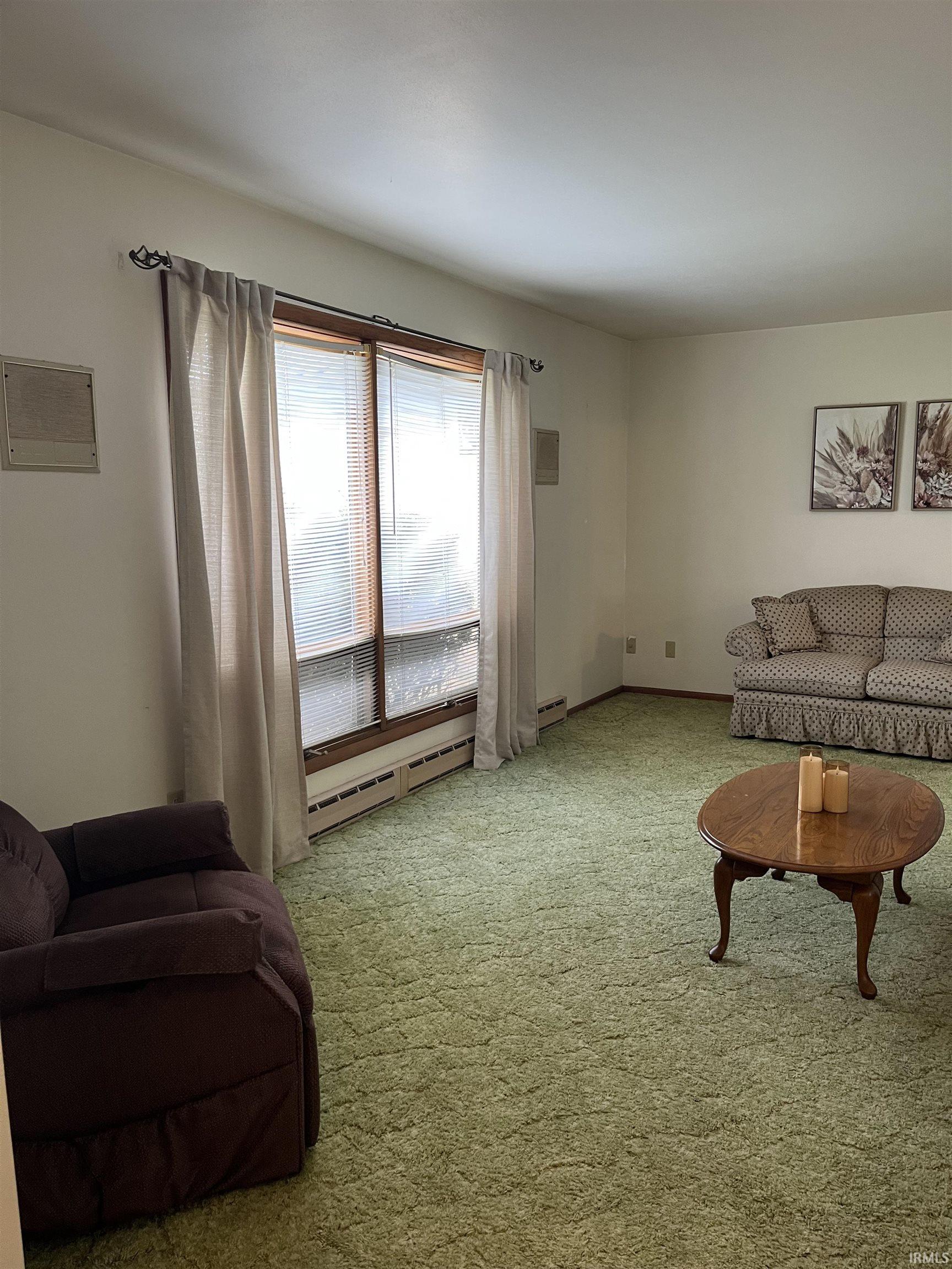 Living area featuring a large window with horizontal blinds, wall-mounted air vents, and perimeter baseboard heating