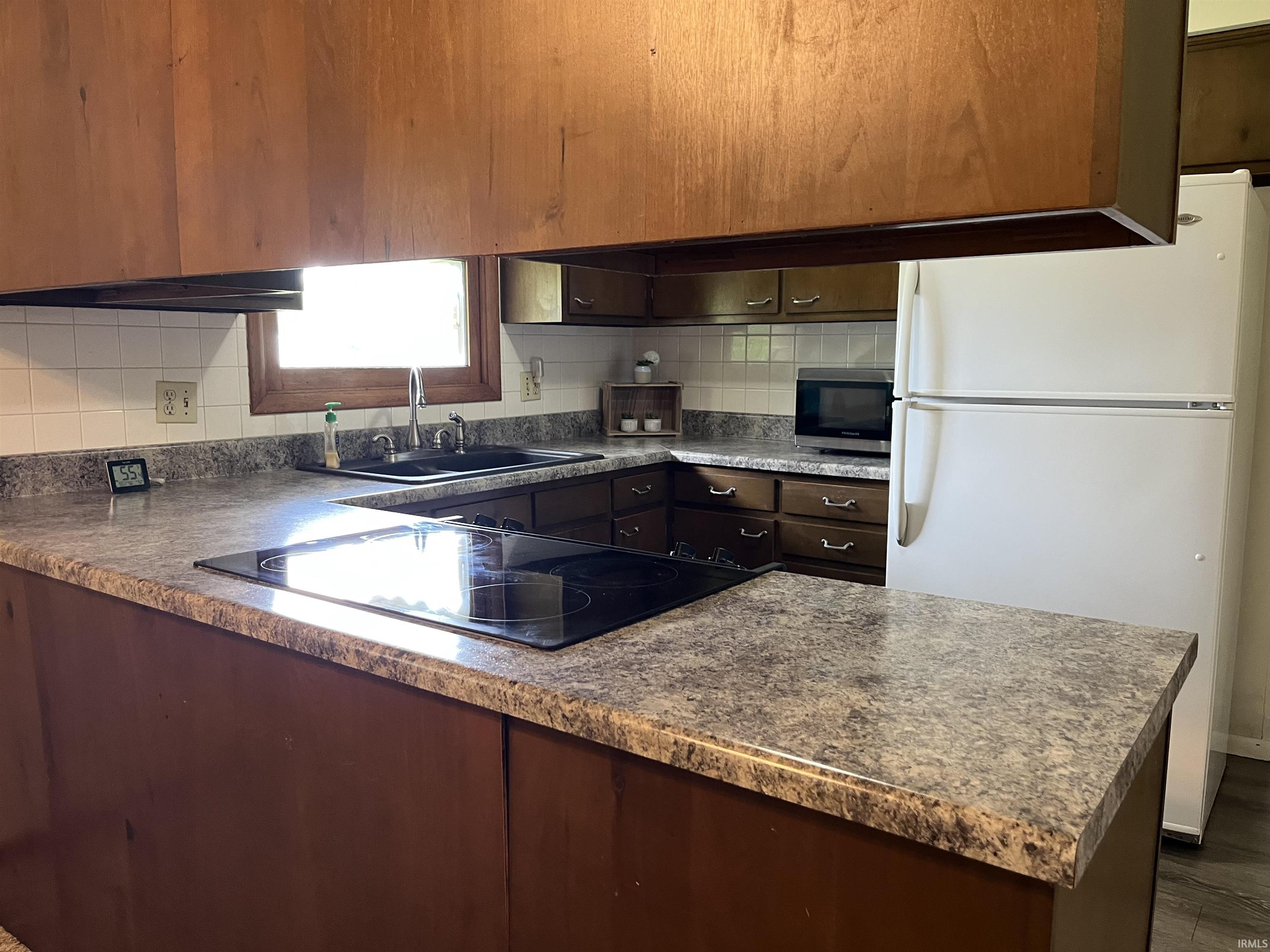 Kitchen featuring laminate countertops, dark wood-finish cabinetry, a built-in electric cooktop, a stainless steel sink with a gooseneck faucet, and a white tile backsplash