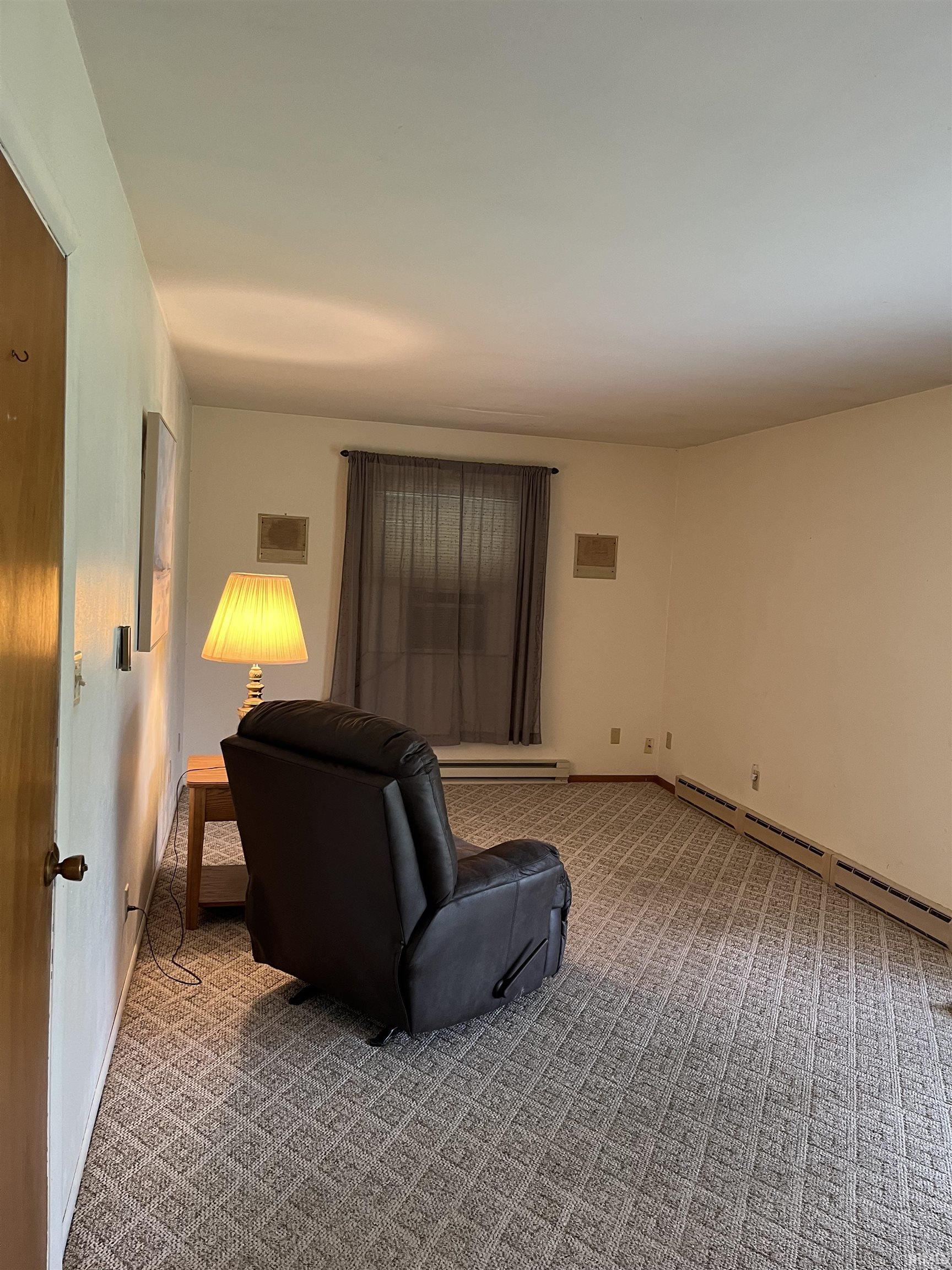 Spacious room featuring patterned carpet, light-colored walls, a window with dark drapes, and baseboard heating units