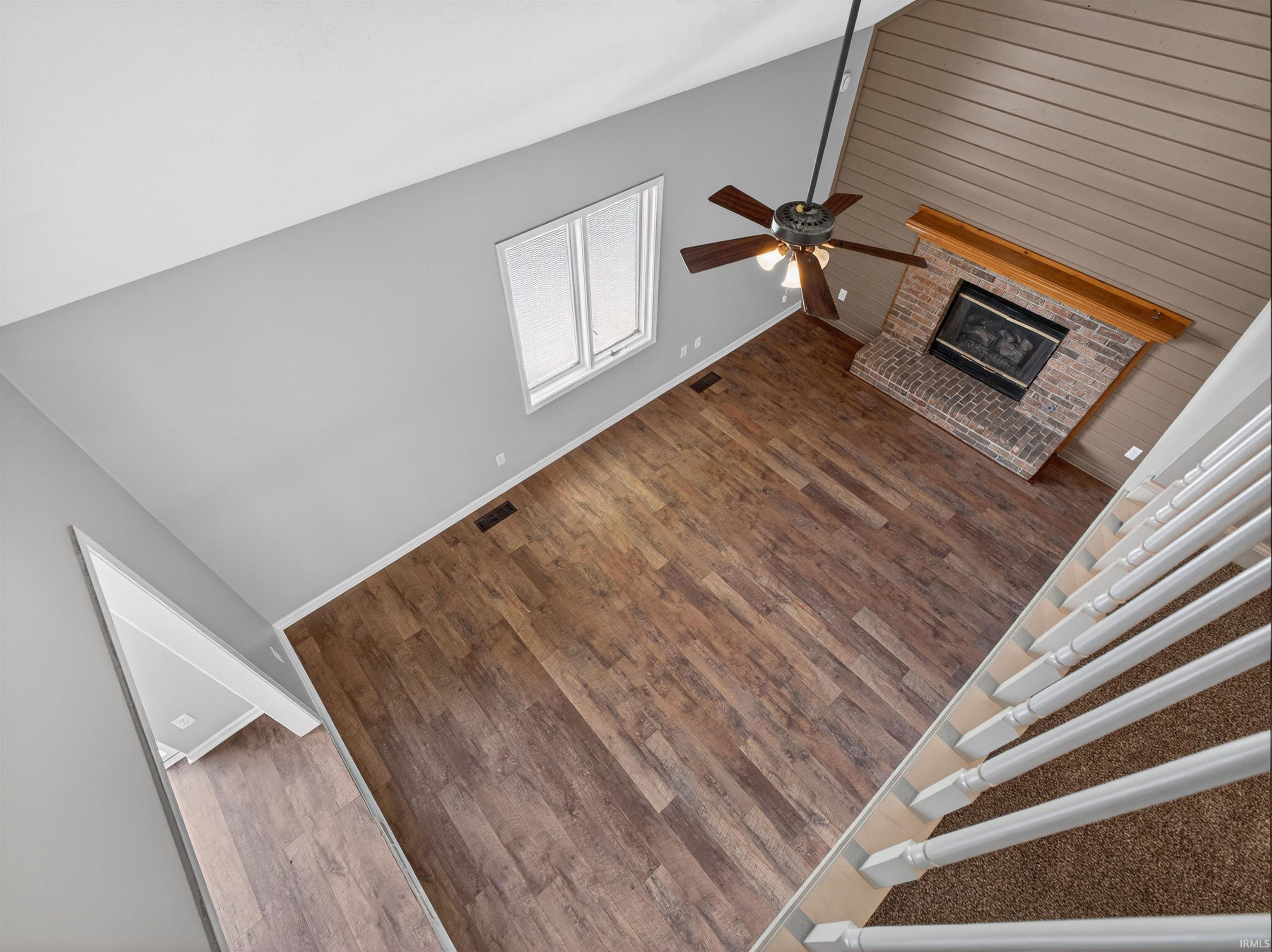 Spacious living area featuring high vaulted ceilings, wood-finish flooring, a brick fireplace with a wood mantel, a ceiling fan with integrated lighting, and a white-trimmed window