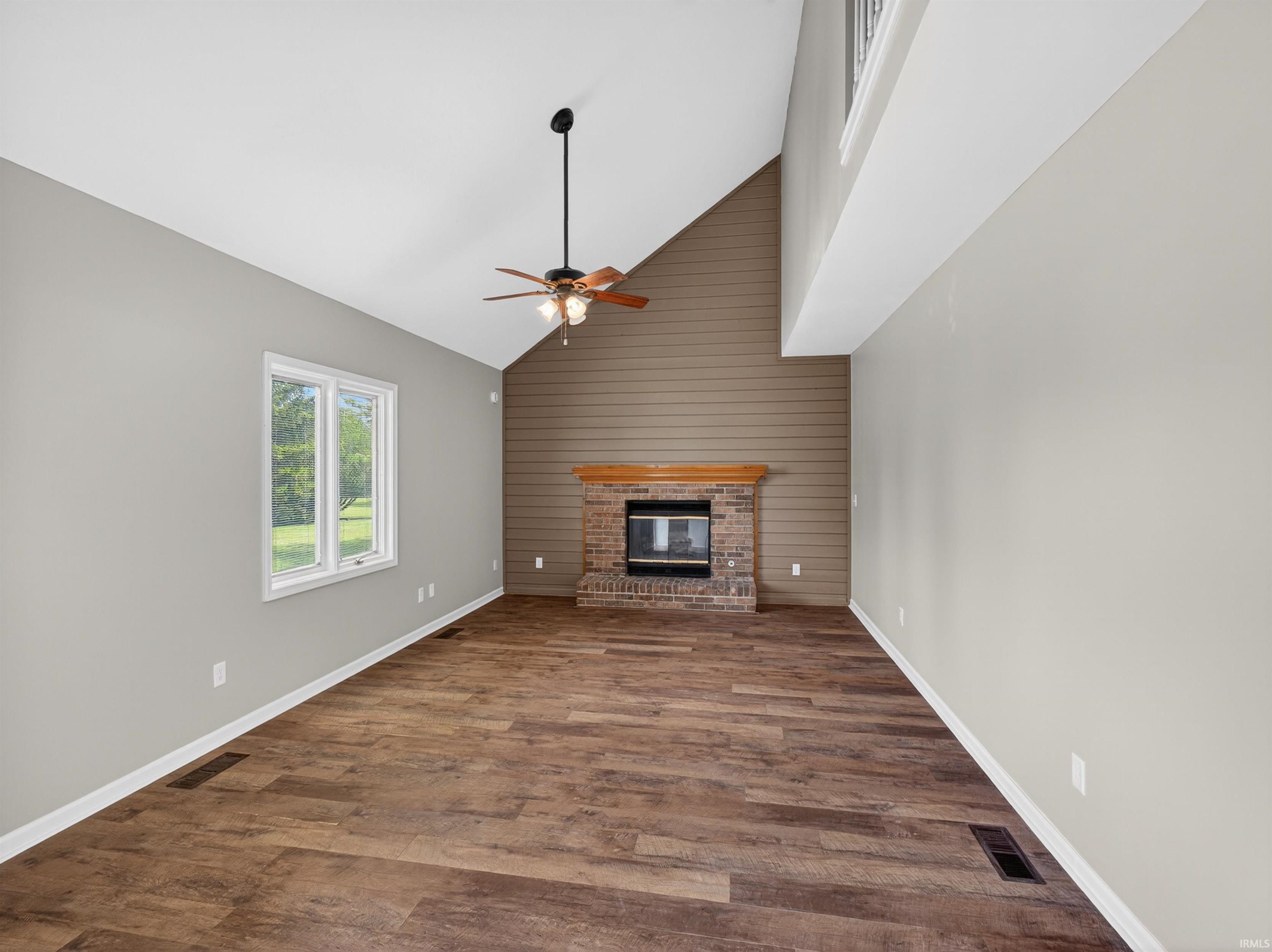 Spacious living area featuring vaulted ceilings, wood-finish flooring, and a brick fireplace with a wood mantel