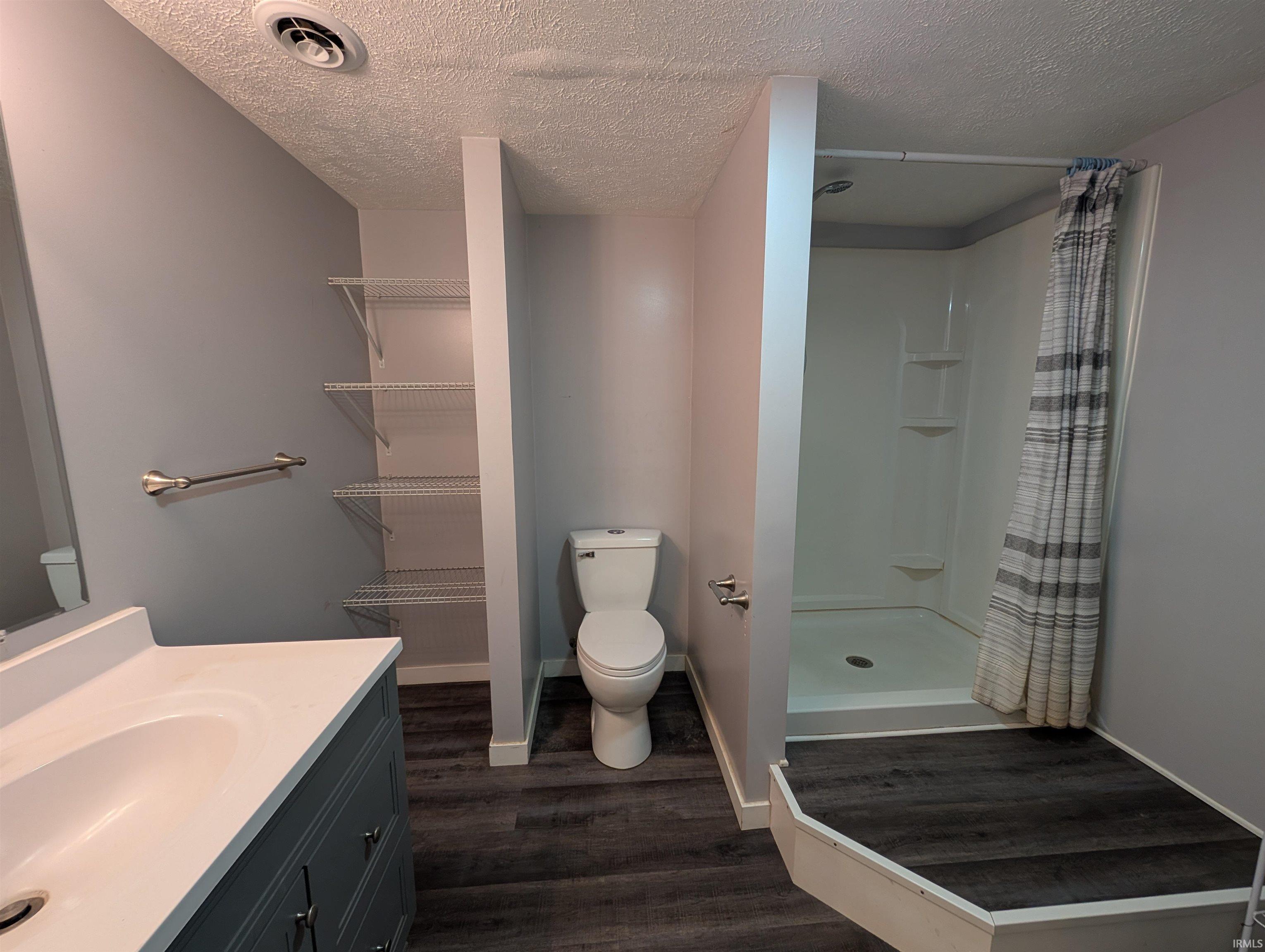 Bathroom featuring a single vanity with a white countertop, dark wood-finish flooring, a white toilet, and a stand-up shower with a white enclosure