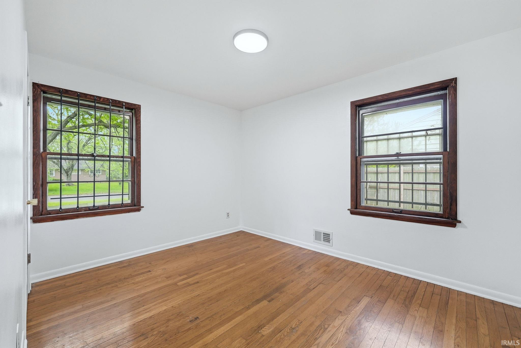 Room featuring hardwood flooring, white walls, two windows with dark wood trim, and a flush mount ceiling light