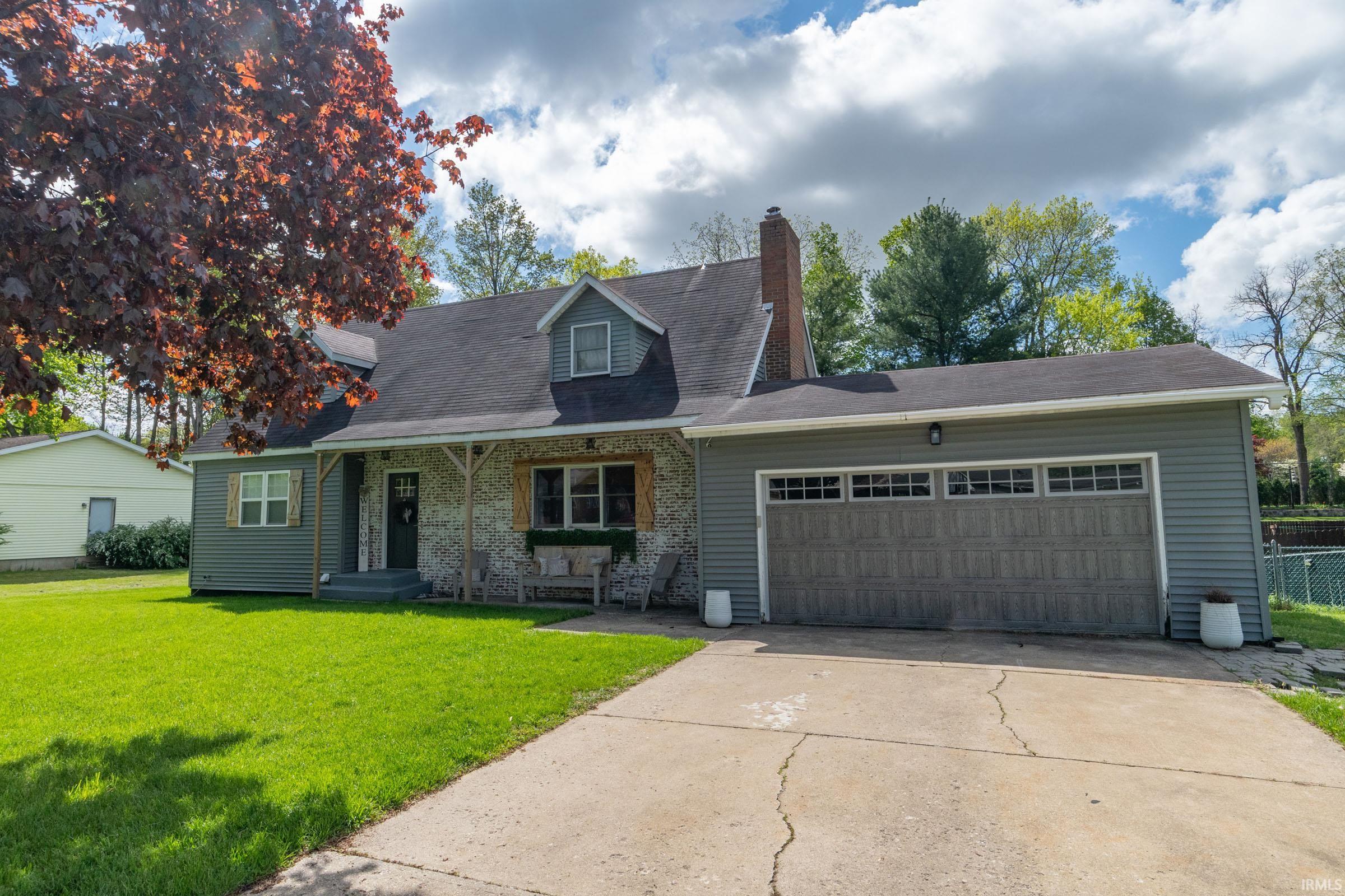 Exterior featuring gray siding, a prominent two-car garage with transom windows, a covered front porch with stone facade, a dormer window, and a dark shingle roof