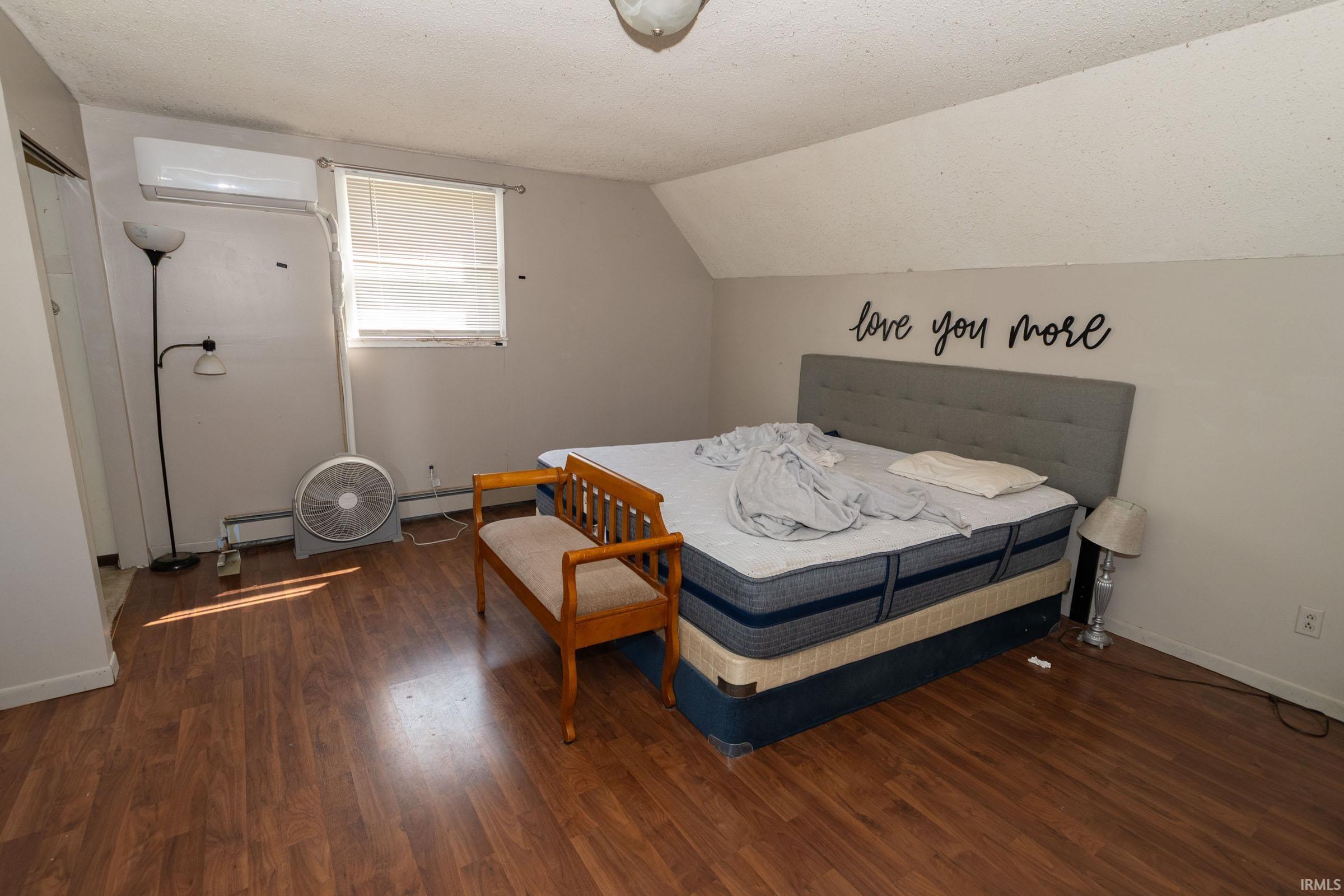 Bedroom space featuring warm wood-finish flooring, a window offering natural illumination, and a wall-mounted mini-split system