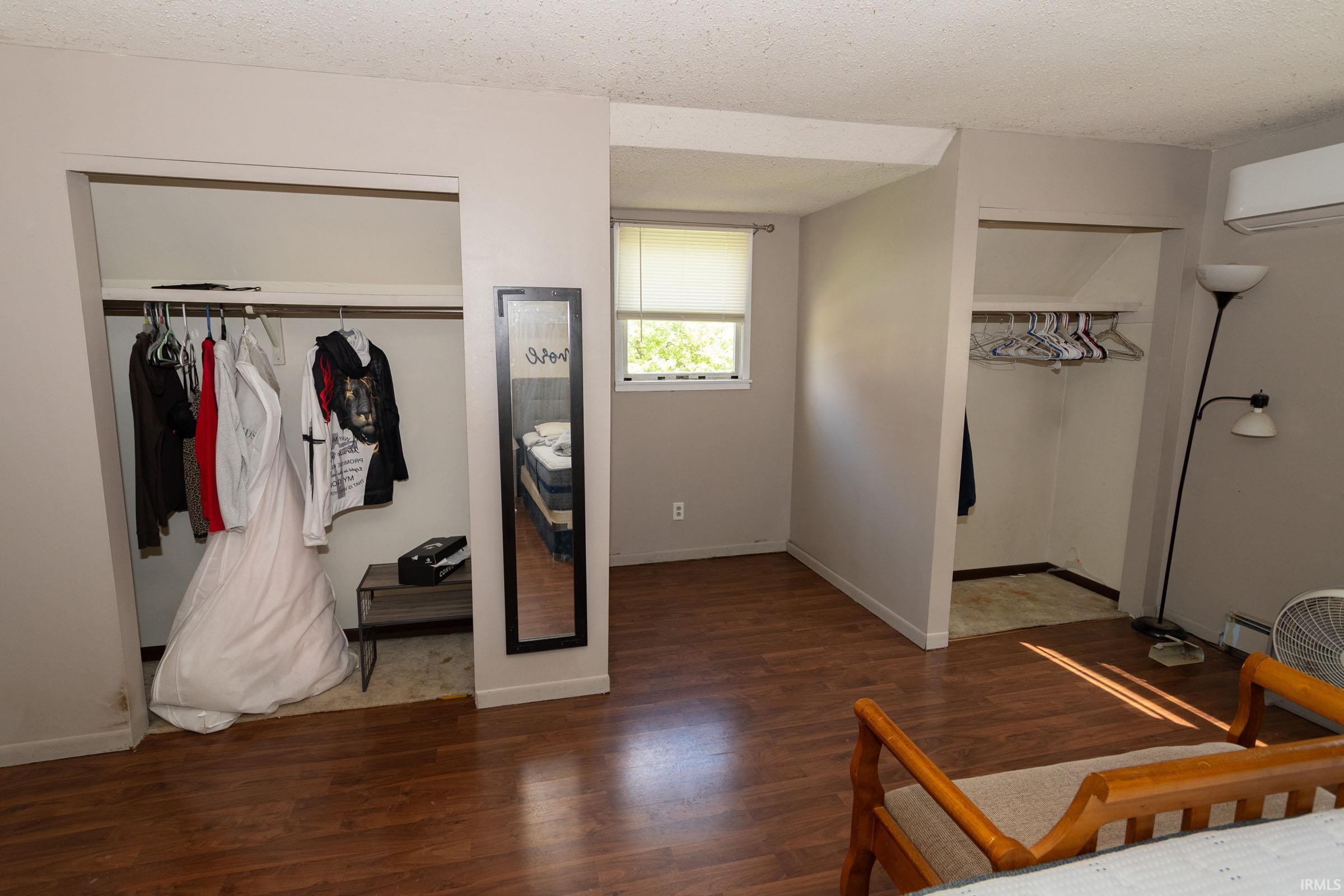 Spacious room featuring rich wood-finish flooring, two open closet areas with hanging rods, a sunlit window with a roller shade, and a wall-mounted air conditioning unit