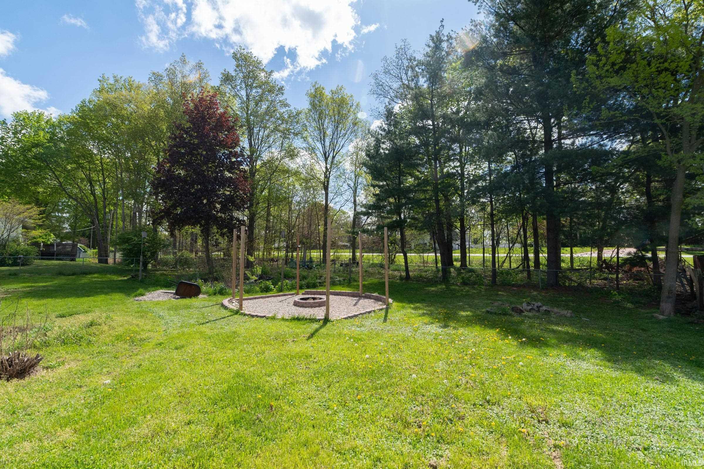 Spacious green lawn with a circular gravel fire pit area, surrounded by mature trees and natural landscaping