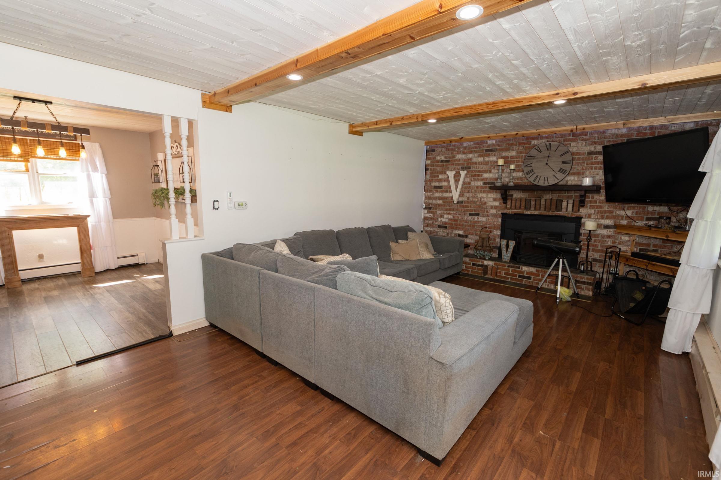 Living area featuring an exposed brick fireplace with a wood mantel, wood-finish flooring, and a white plank ceiling with exposed wood beams and recessed lighting