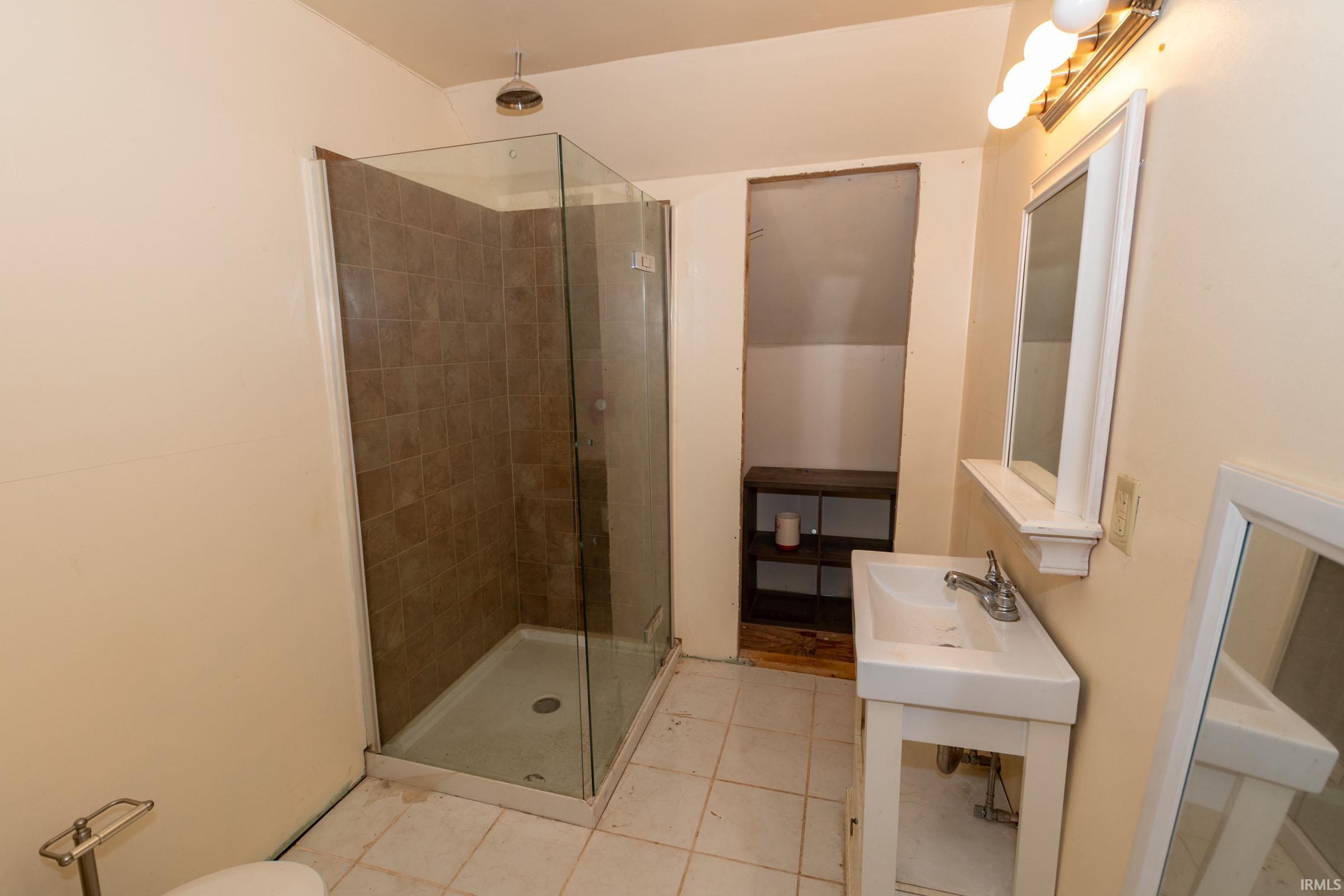 Glass-enclosed shower with tiled interior and rainfall showerhead, white porcelain vanity with chrome fixtures, and built-in shelving