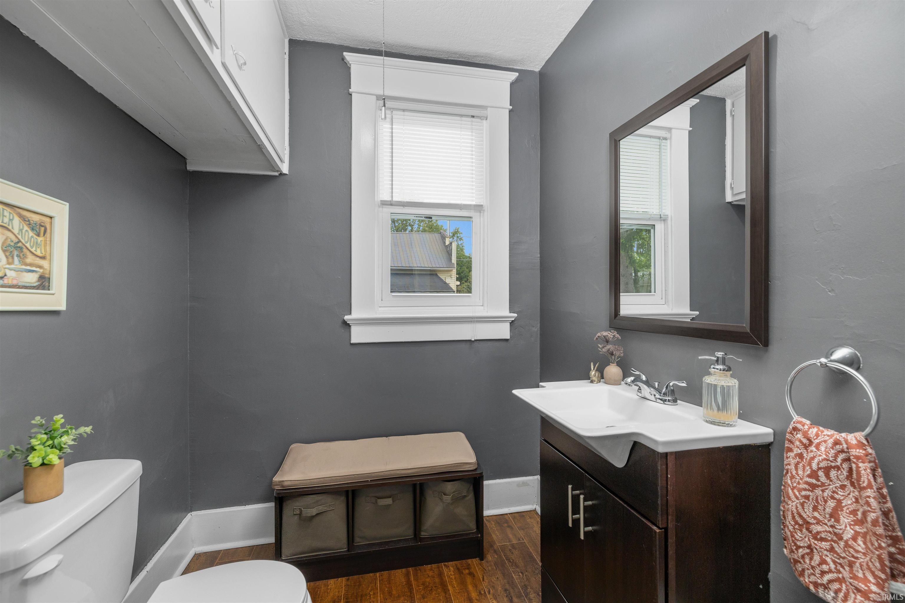 Bathroom featuring a dark wood-finish vanity with an integrated sink, framed mirror, wood-finish flooring, and a window with white trim